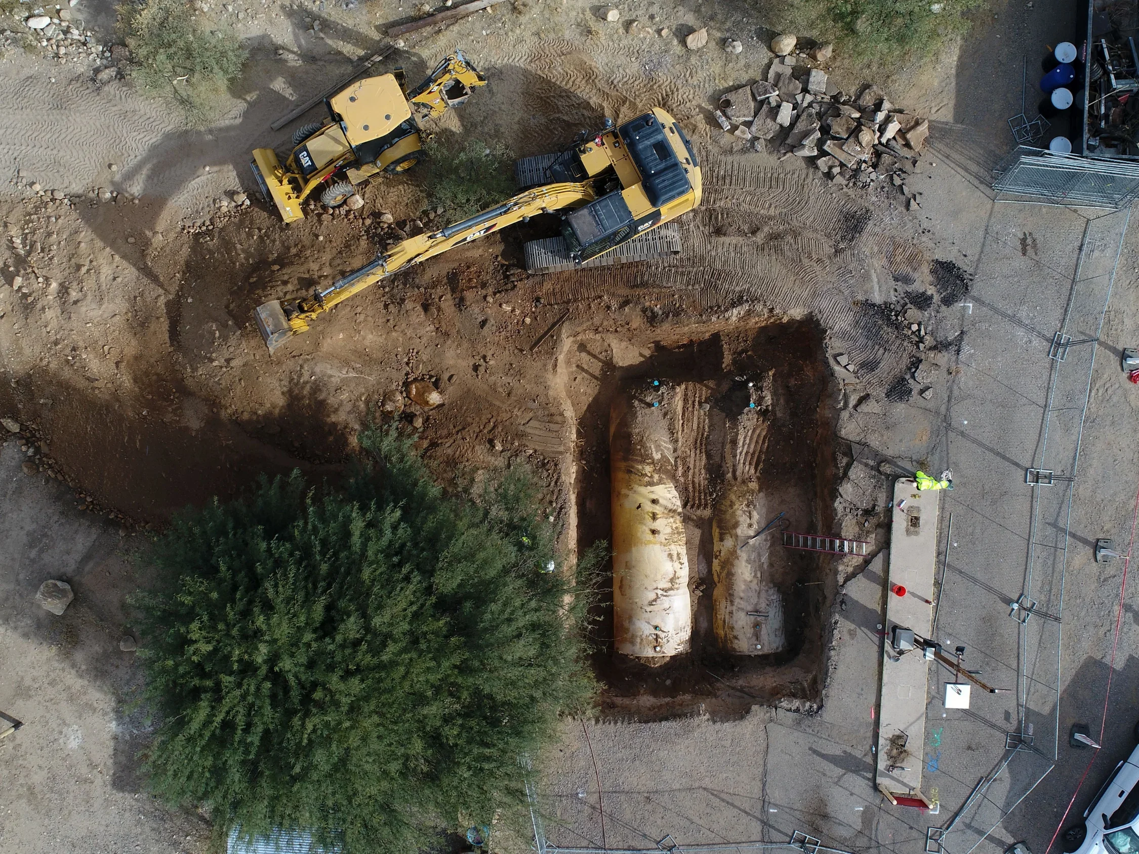 Aerial view of a construction site with a large excavator and bulldozer moving dirt, rocks, and pipe underground, surrounded by construction barriers and equipment.