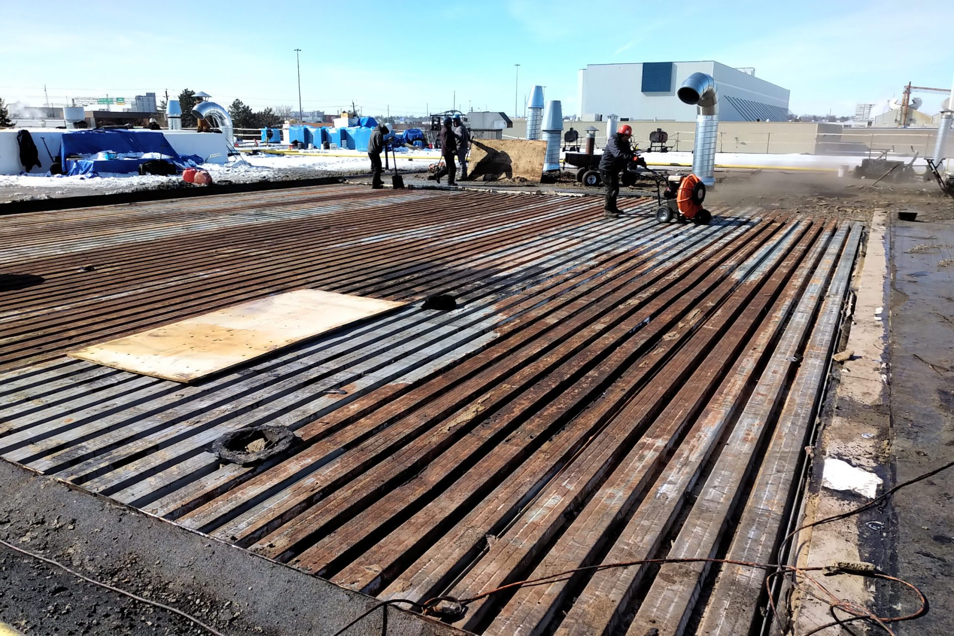 Construction workers are working on a rooftop, installing or repairing the metal decking. The area is outdoors with equipment, pipes, and other construction materials visible in the background.