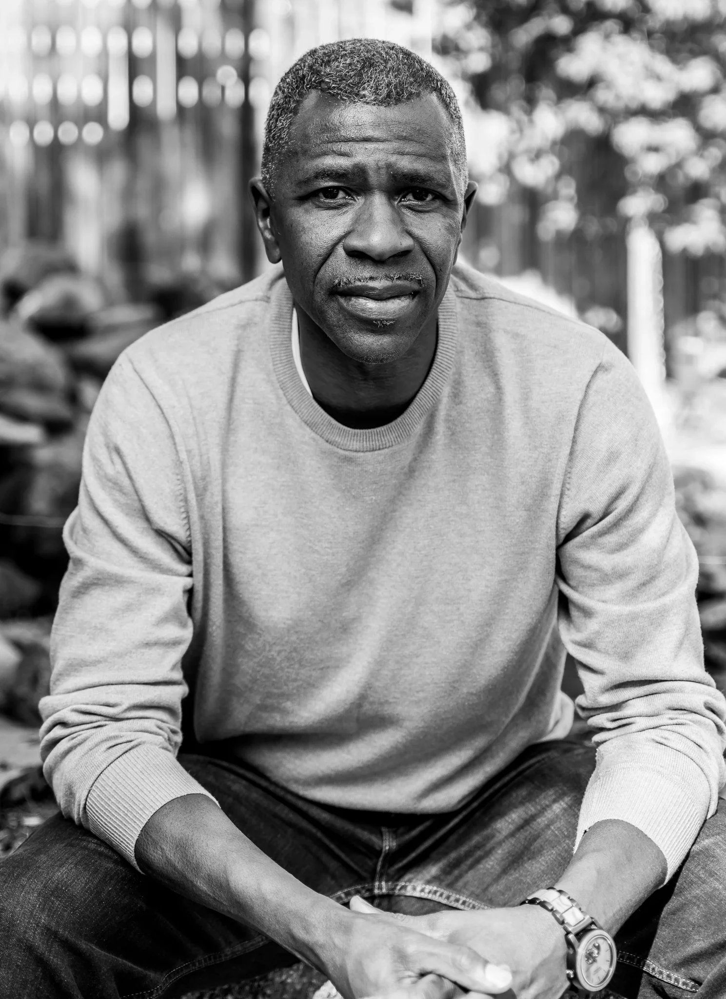 A black and white photo of a middle-aged man with short hair, wearing a light-colored sweatshirt, sitting outdoors with a fence and trees in the background.