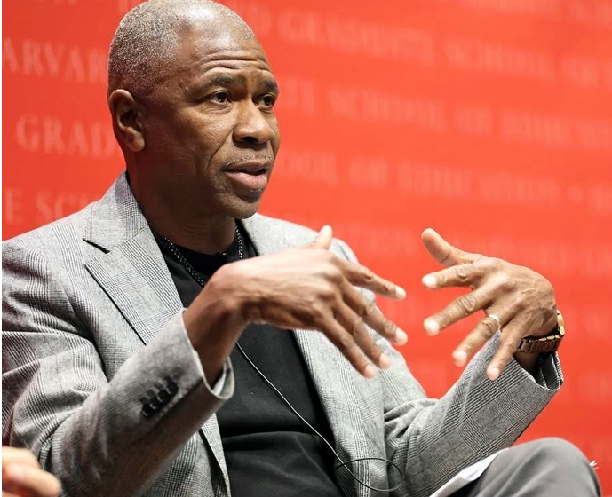 African American man in a gray blazer gesturing with his hands during a discussion or interview against a red background.