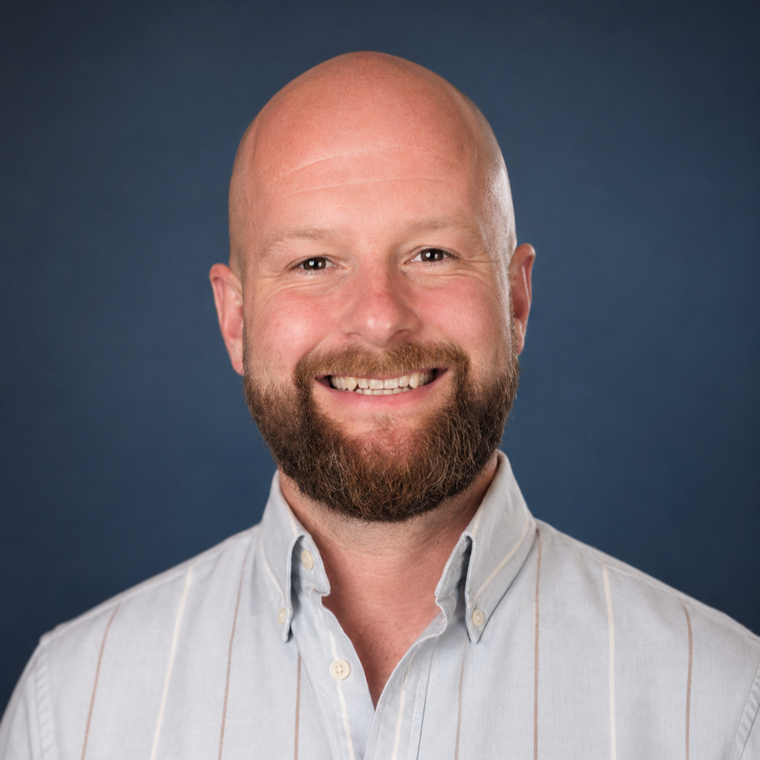 Headshot of a smiling bald man with a beard, wearing a light-colored button-up shirt, against a dark blue background.