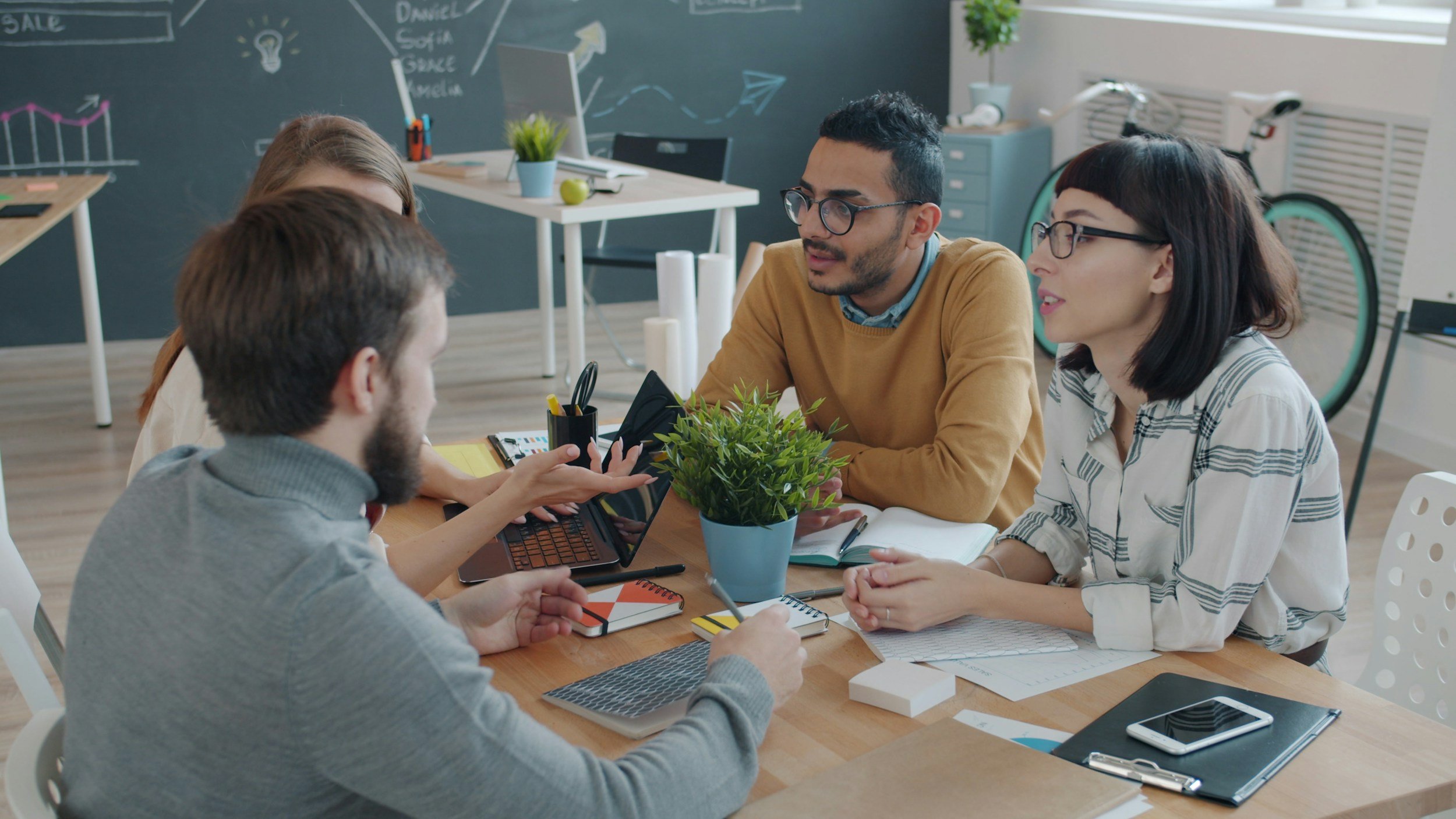 Group of five young professionals in a meeting, discussing around a wooden table with notebooks, a laptop, pens, and a potted plant, in a bright office with a chalkboard wall in the background.