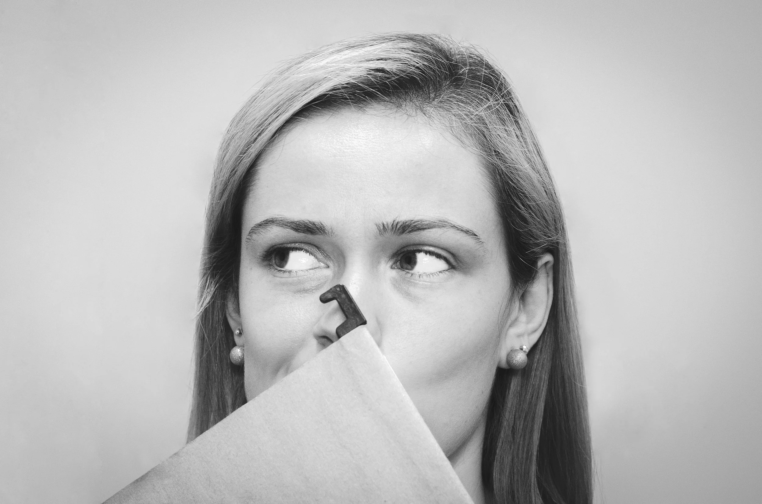 Black and white photo of a woman with light-colored hair, wearing earrings, holding a piece of paper or card in front of her face, partially obscuring it, with her eyes looking to the side.