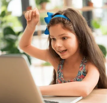 A young girl with a blue bow in her hair smiling and raising her fist while looking at a laptop.