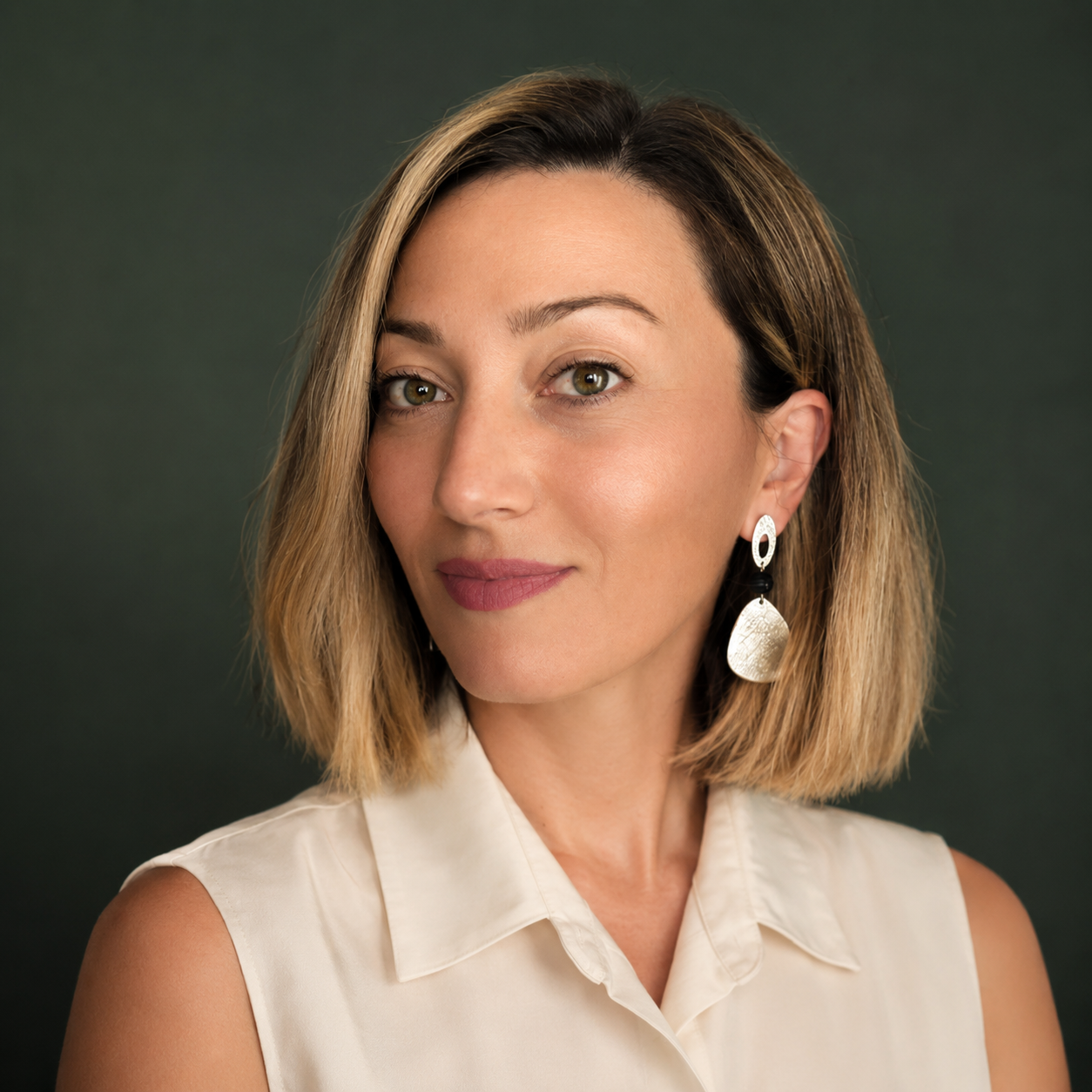 A woman with shoulder-length blonde hair wearing a cream sleeveless top and statement earrings, looking at the camera with a slight smile against a dark background.