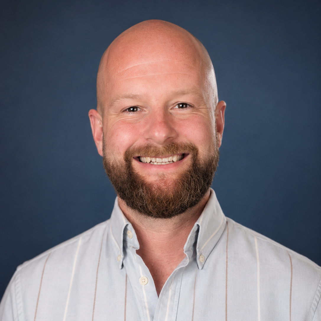 Headshot of a smiling bald man with a beard, wearing a light-colored striped button-up shirt against a dark blue background.