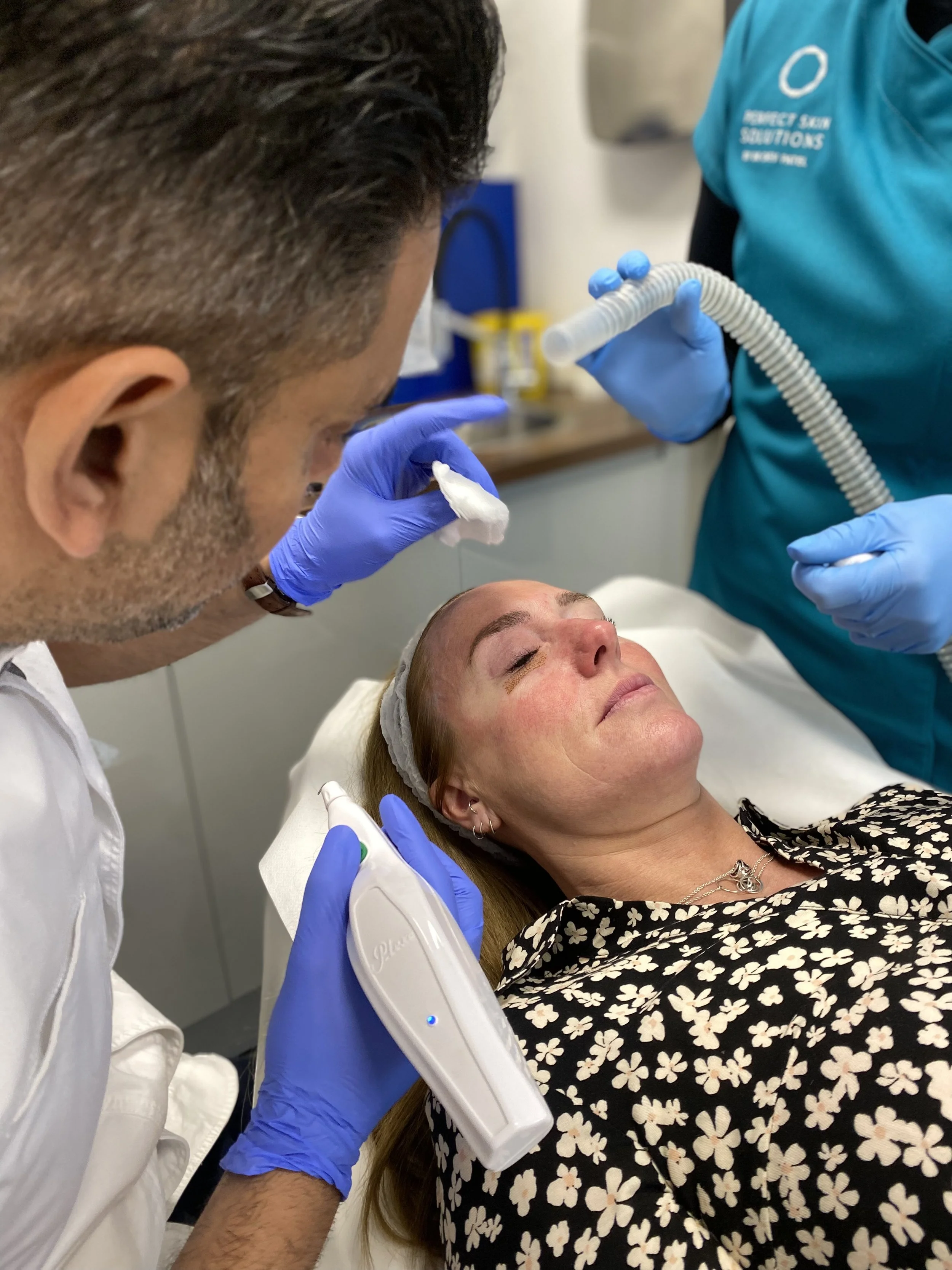 A woman lying on a bed receives a cosmetic or medical treatment to her face from a professional, who is approximately in his thirties or forties and wears blue gloves. The professional holds a tool and appears to be working on her face. The woman has closed eyes and a relaxed expression.