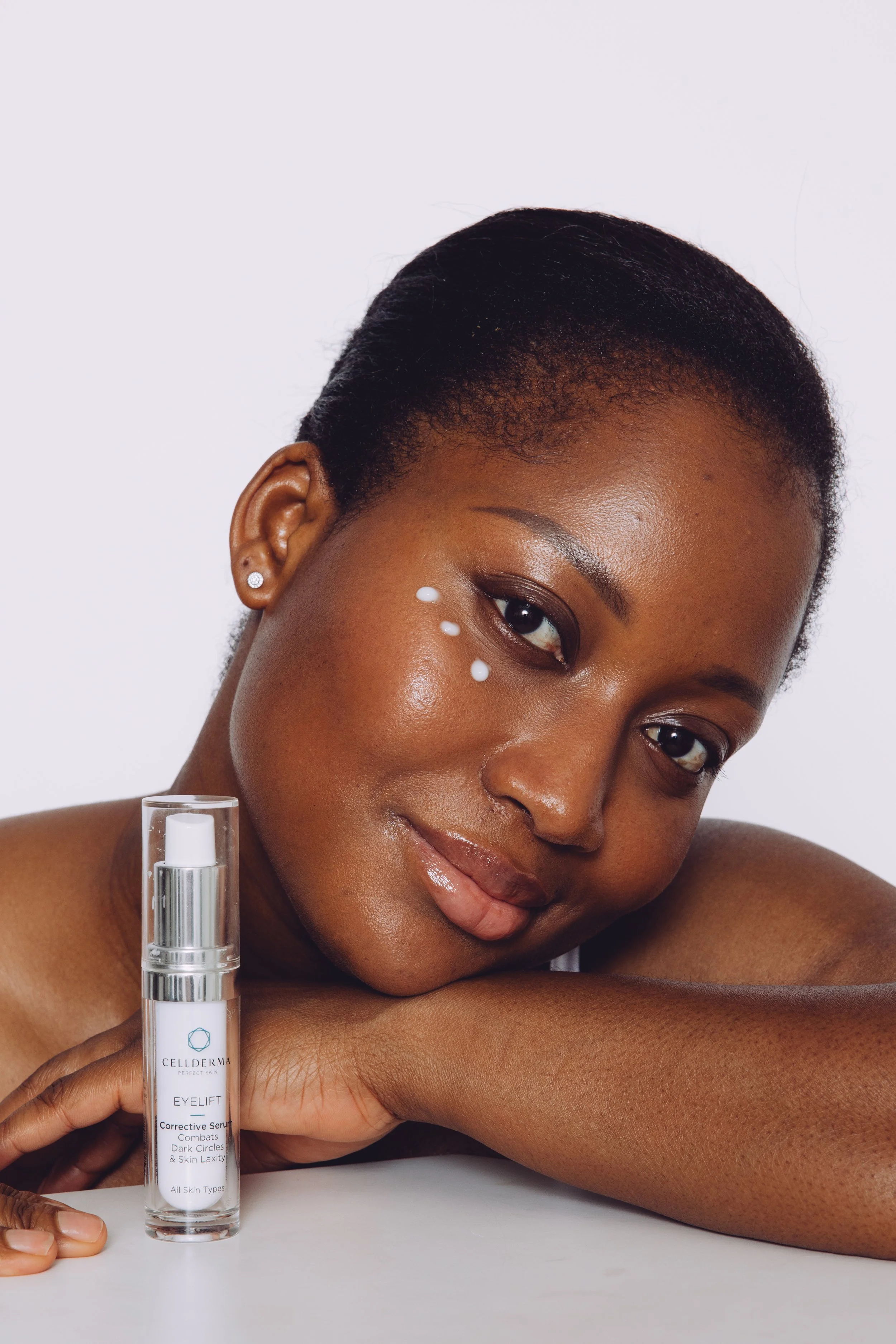 Close-up of a woman with dark skin and short black hair with hair product dots on her face, resting her face on her crossed arms in front of a white background. She has a small earring and is holding a clear spray bottle labeled Cellderma Eyelift in her right hand.