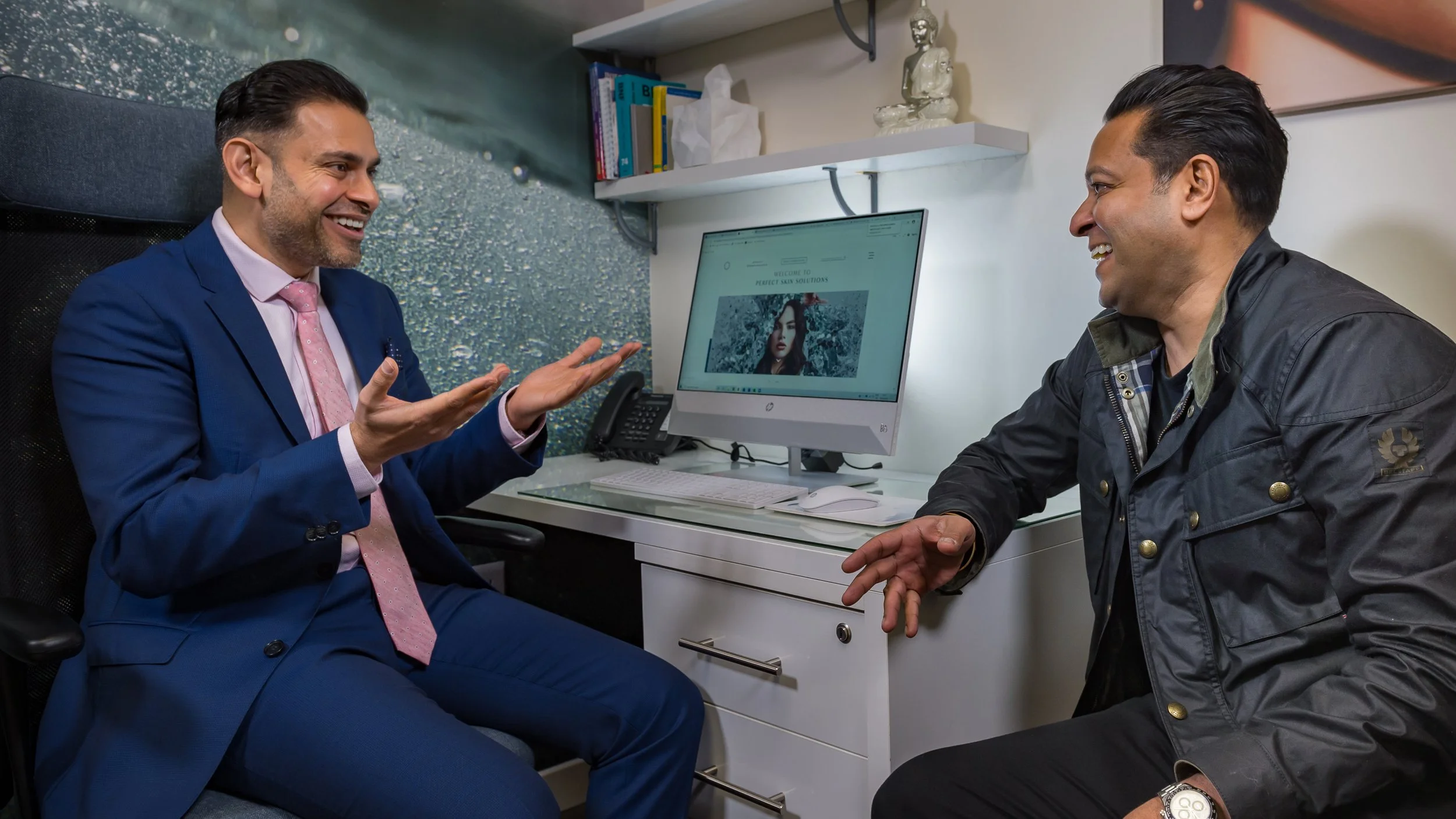 Two men are sitting in an office, engaging in conversation and smiling. One man, dressed in a blue suit and pink tie, is gesturing with his hands. The other man, in a black jacket with gold buttons, is smiling and listening. There is a computer monitor on the desk displaying a webpage, and shelves with books and decorative items in the background.