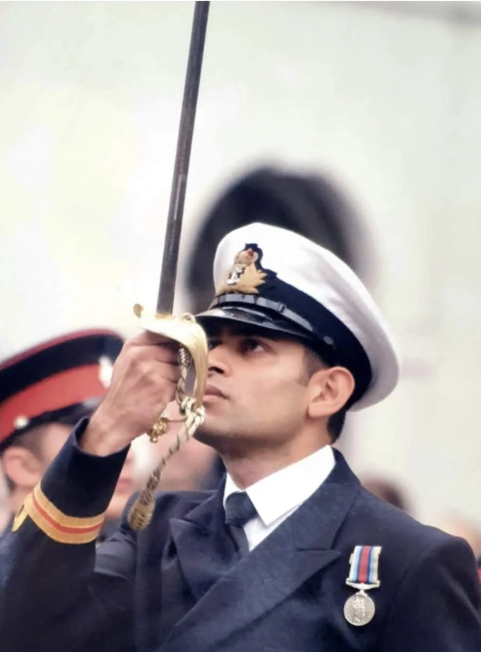 A uniformed officer in a navy suit and white cap with a badge, holding a sword aloft during a formal ceremony or parade.