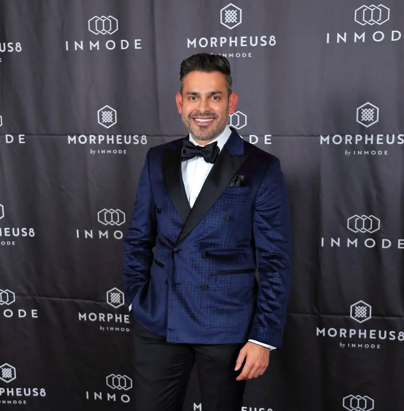 Man in a blue tuxedo with black lapels and a black bow tie, standing in front of a black backdrop with white logos and text for 'INMODE' and 'MORPHEUS8.'
