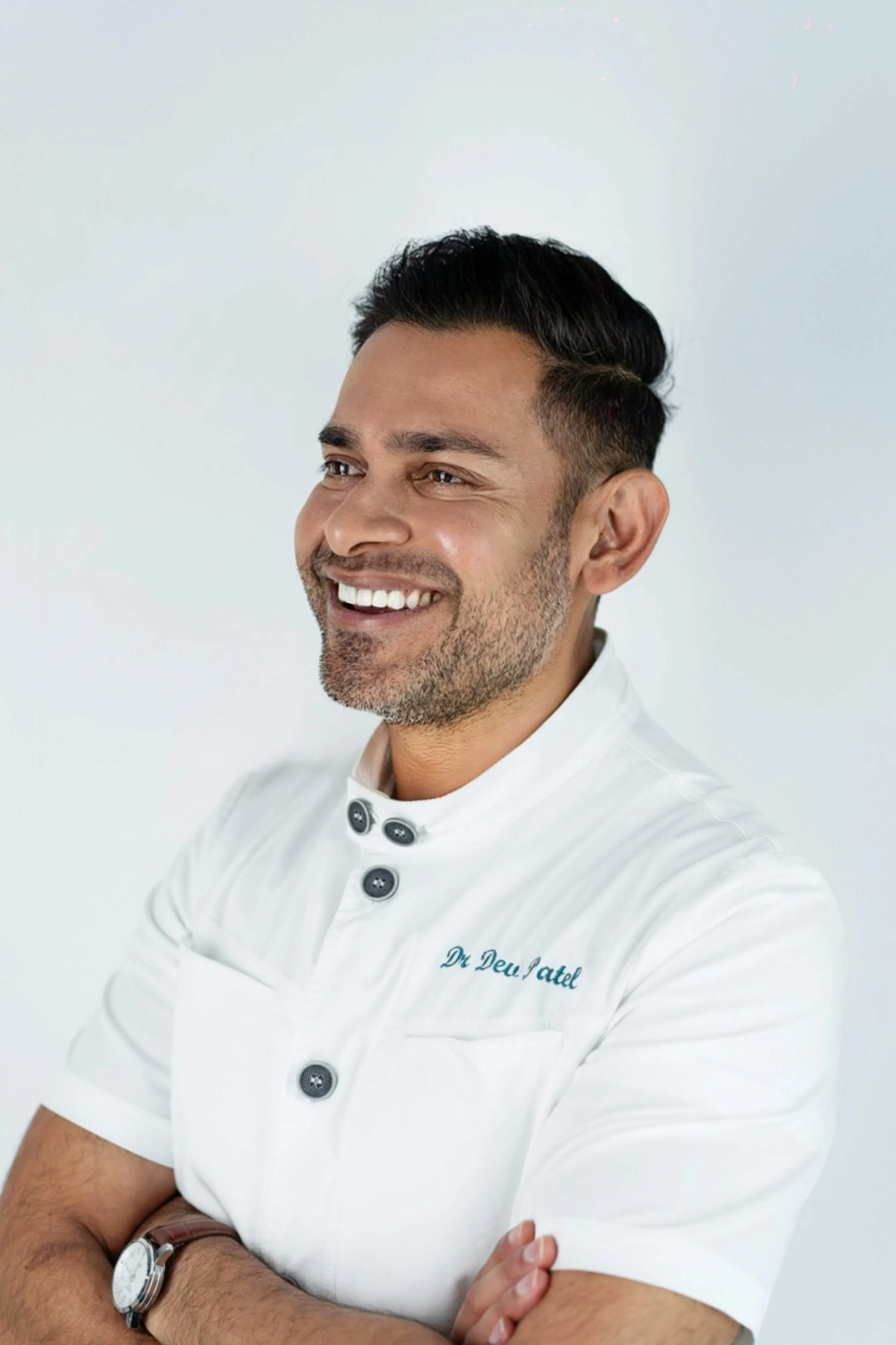 Smiling man with dark hair, wearing a white chef's coat with name embroidered, arms crossed, wearing a wristwatch, against a plain light background.