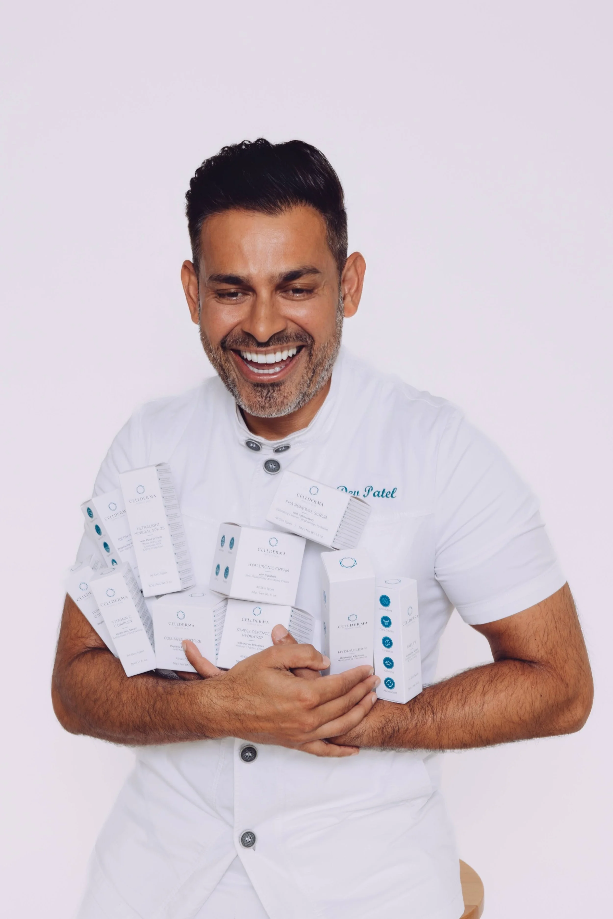 A man in a white shirt smiling while holding various skincare product boxes. The background is plain white.