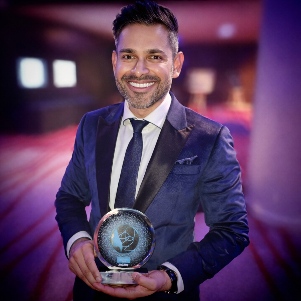 A man in a dark blue tuxedo holding an award with a glass top, standing in a room with colorful lighting and blurred background.