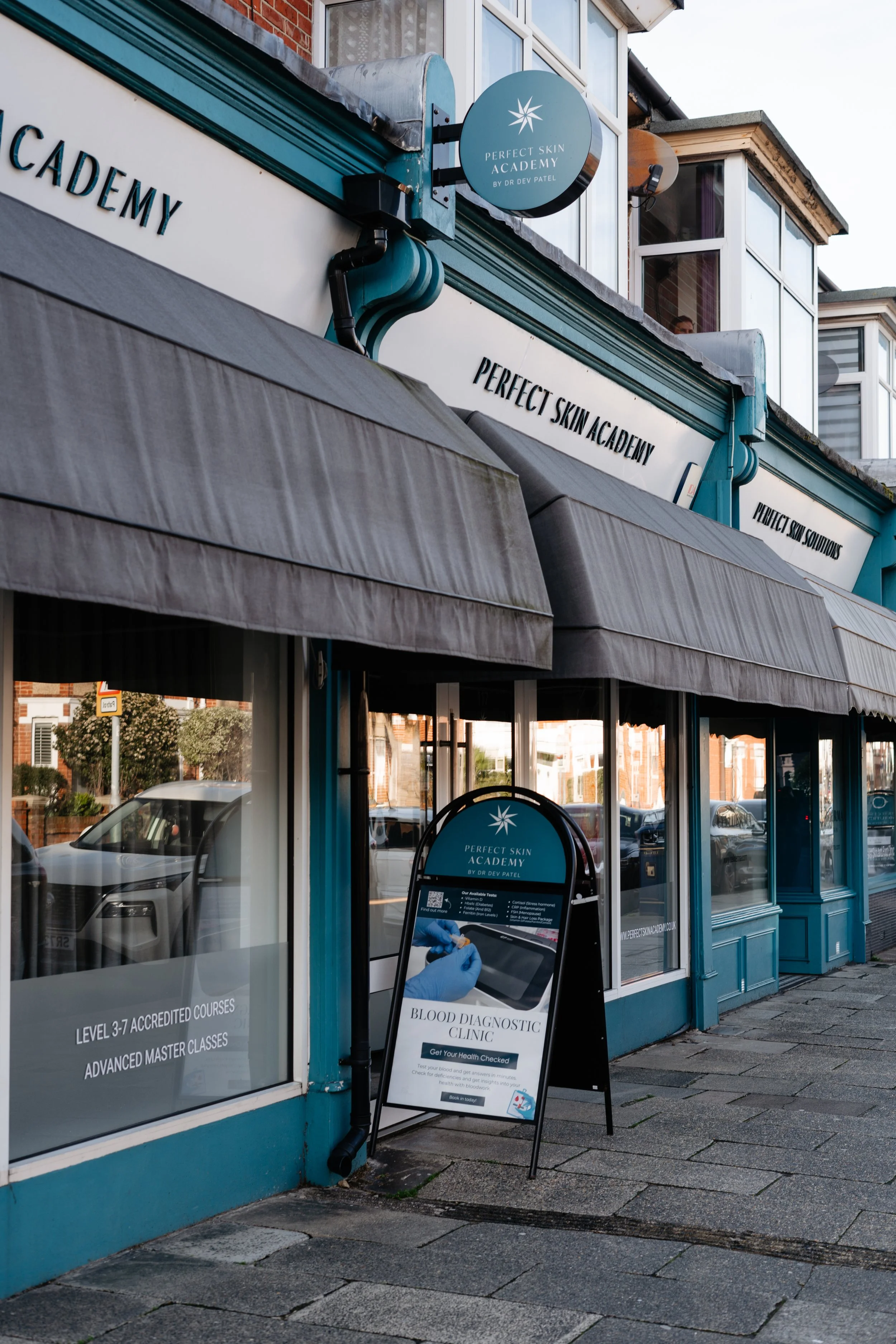 Exterior view of a storefront named 'Perfect Skin Academy' with a signboard outside advertising blood diagnostic clinics and skin courses. The shop has large windows, a grey awning, and a sidewalk in front.