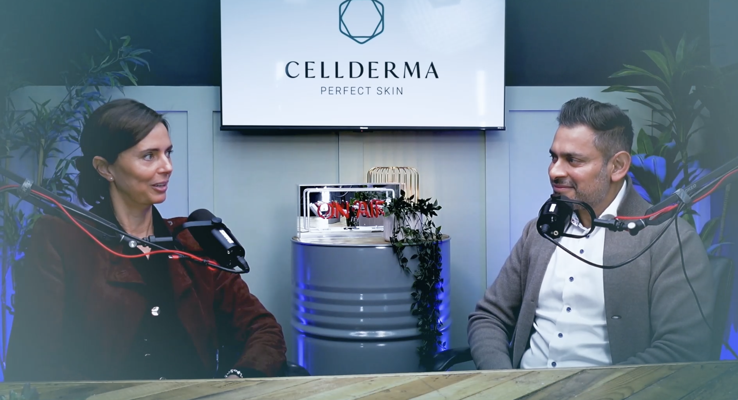 A woman and a man are sitting at a table in a recording studio with microphones, engaged in conversation. Behind them is a large screen displaying the logo and name of Cellderma, a skincare brand, with the slogan "Perfect Skin." The background features green plants and modern decor, and there are small decorative objects on the table.