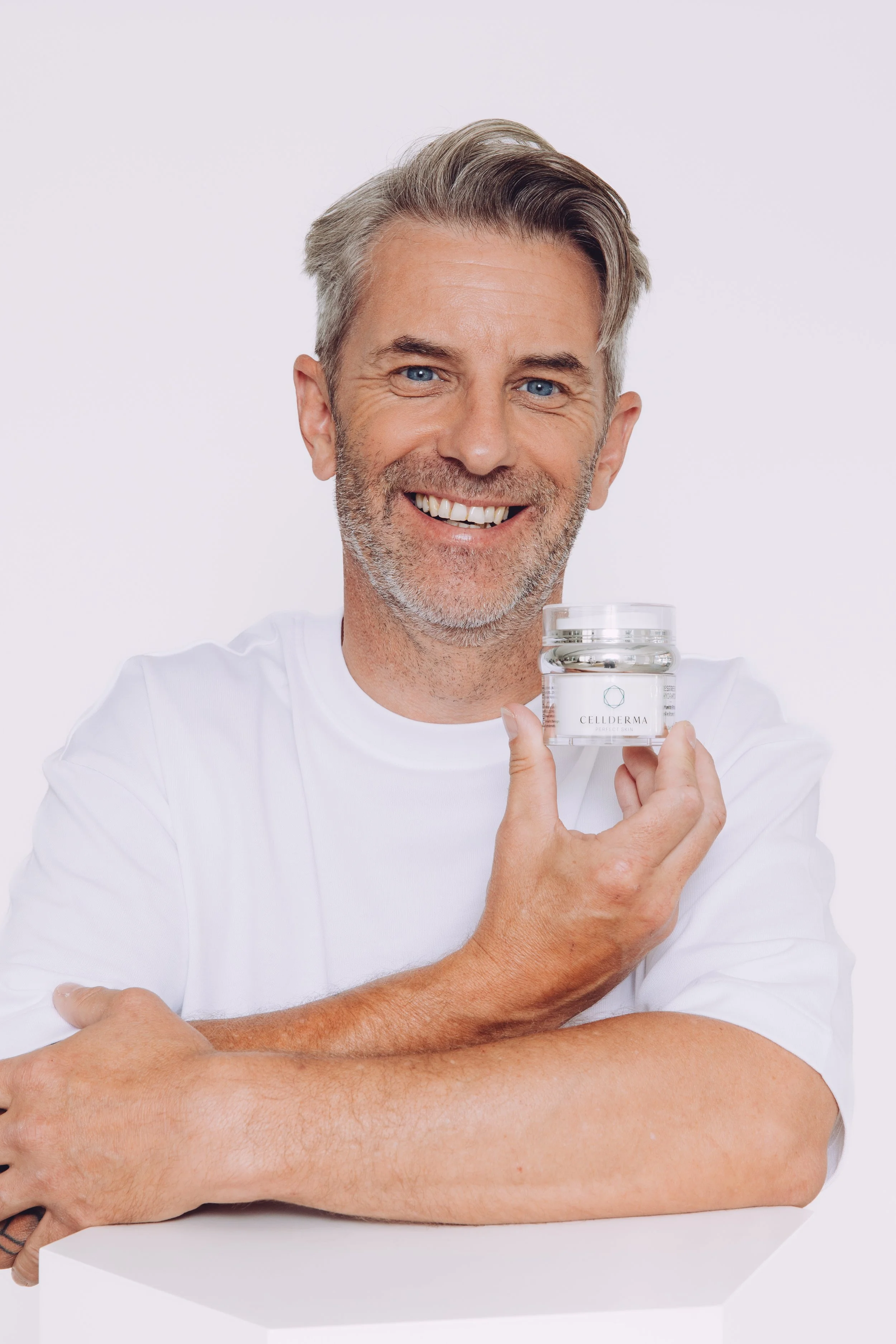 A smiling middle-aged man with gray hair and beard, wearing a white shirt, holding a jar of skincare product labeled 'CELDERMA' against a white background.