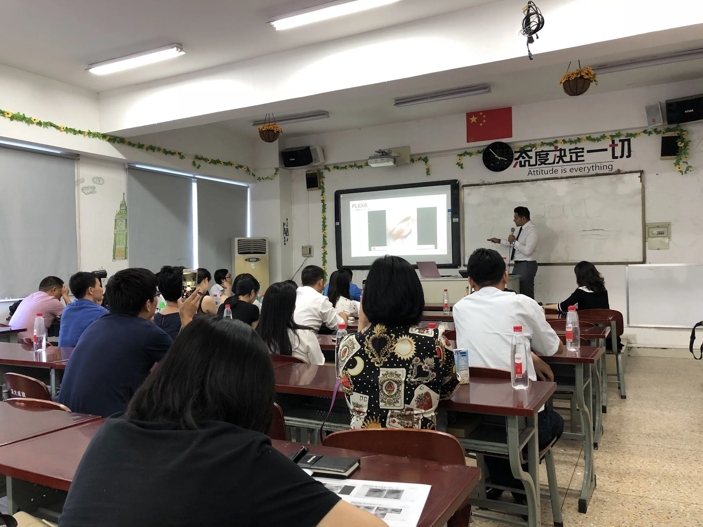 A classroom filled with students sitting at desks, listening to a male presenter standing next to a whiteboard with a projector screen displaying a presentation. Decorations include a Chinese flag and a banner with Chinese characters and the phrase 'Attitude is everything.'