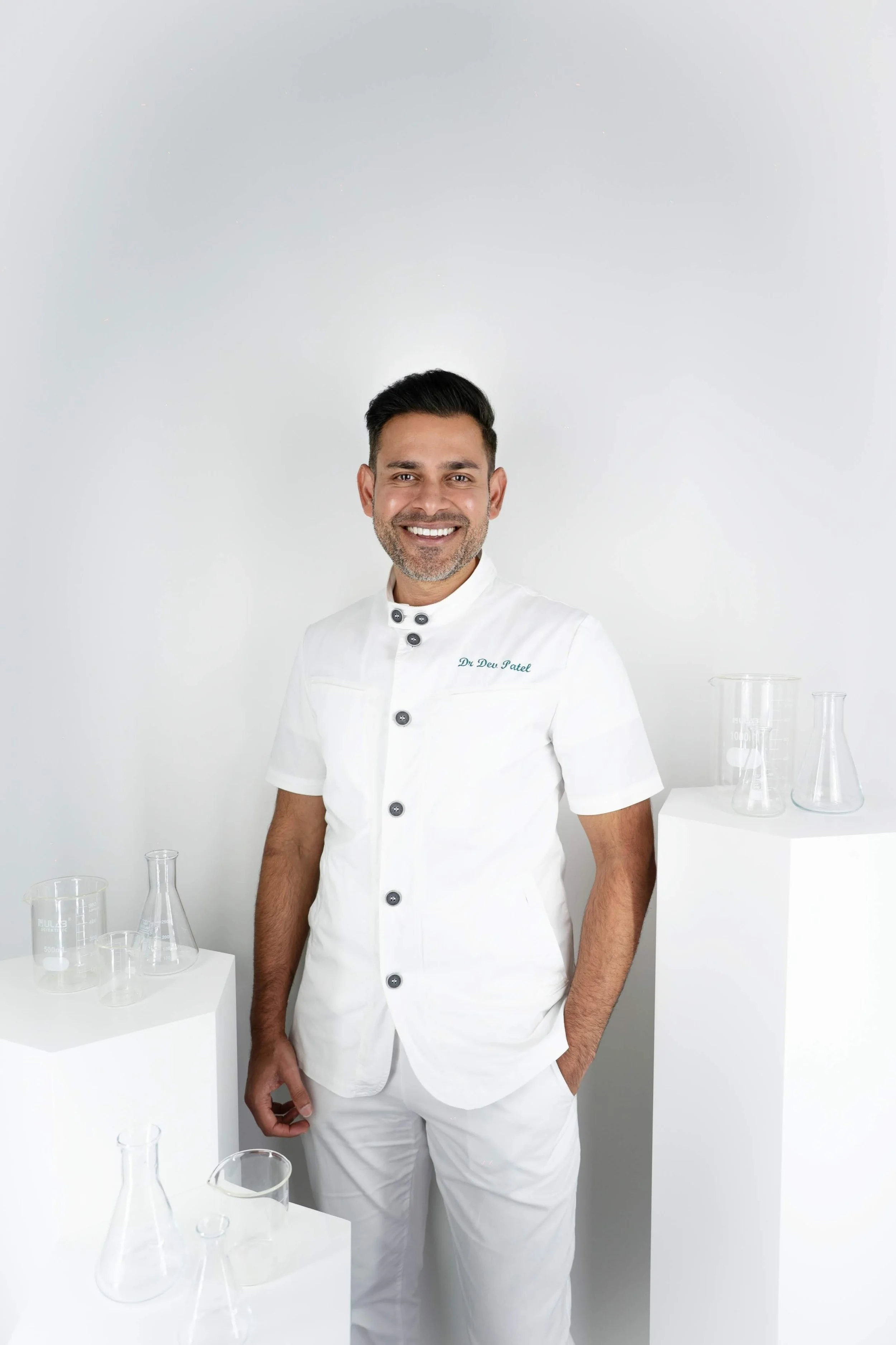 A man in a white chef uniform standing in a bright white room, smiling. There are lab glassware and beakers on white pedestals around him.