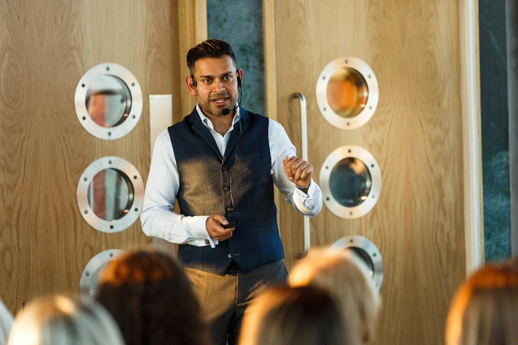 A man wearing a headset with a microphone giving a presentation or speech in front of a wooden wall with circular windows, with an audience visible in the foreground.