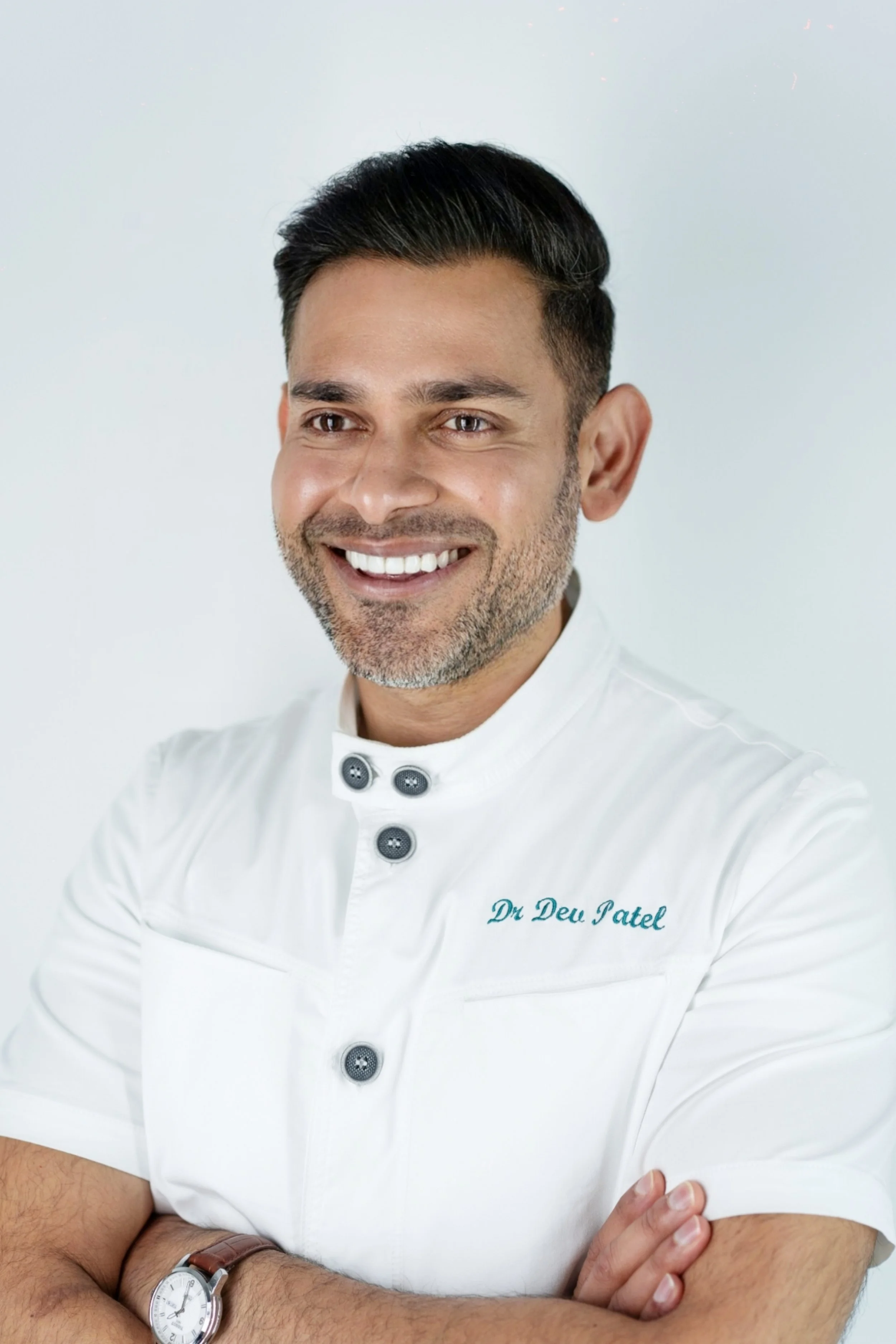 A smiling man in a white medical uniform with embroidered name 'Dr. Dev Patel' stands with arms crossed, wearing a wristwatch, against a plain white background.