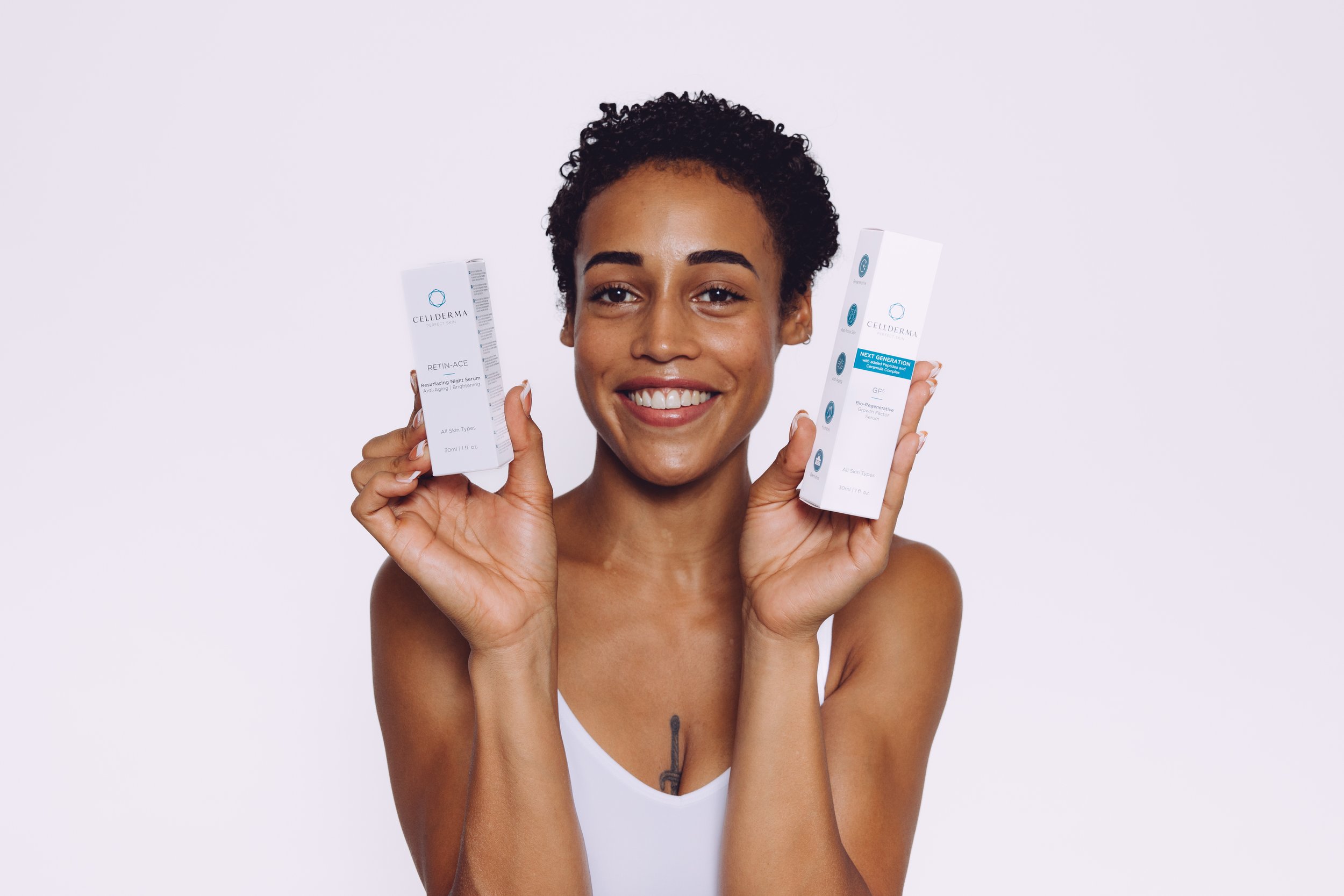 A woman with short curly hair holding two skincare product boxes smiling at the camera against a plain white background.