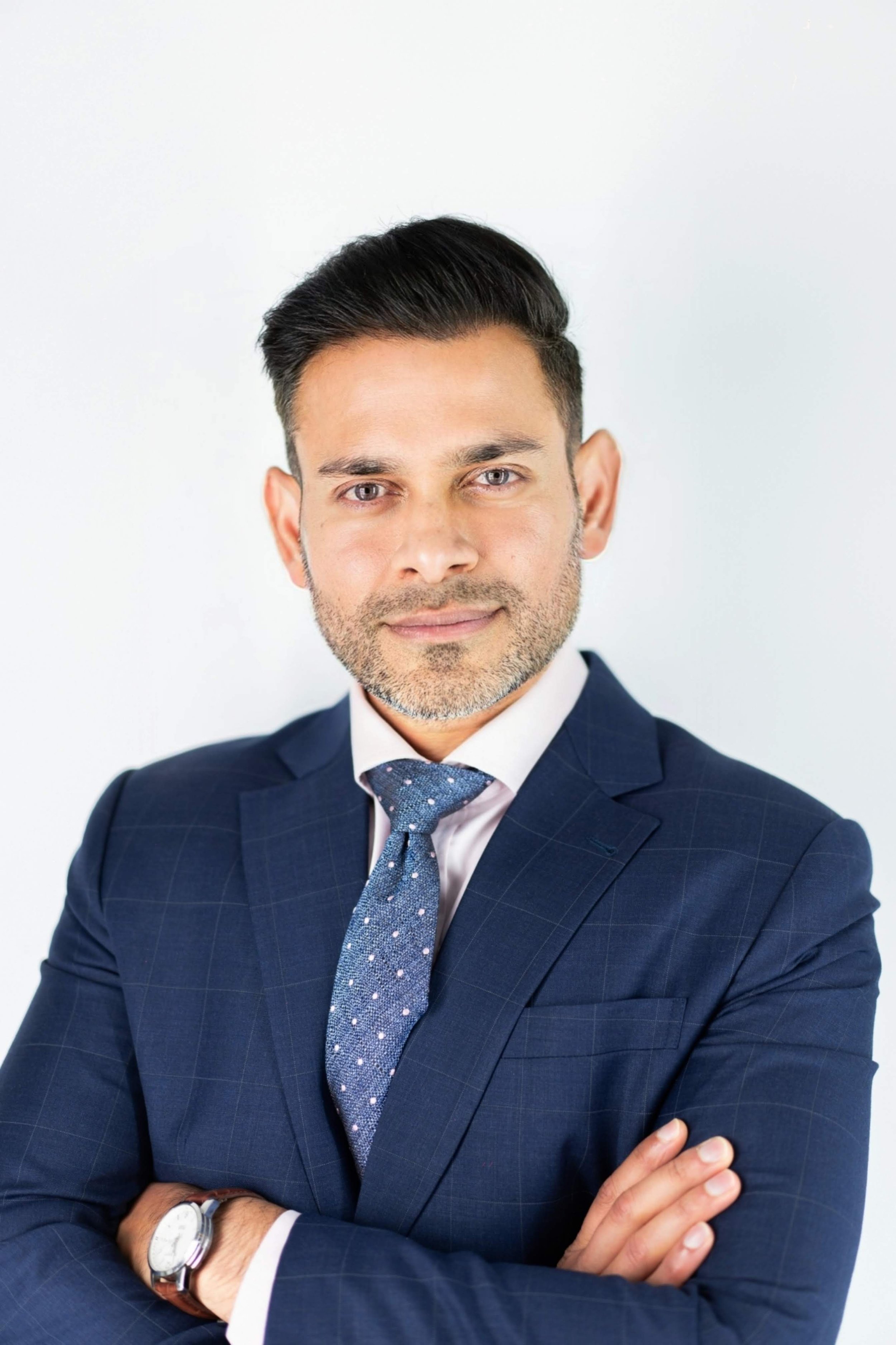 A confident man in a navy blue suit with a light blue tie, crossing his arms, looking at the camera, wearing a watch, against a plain white background.