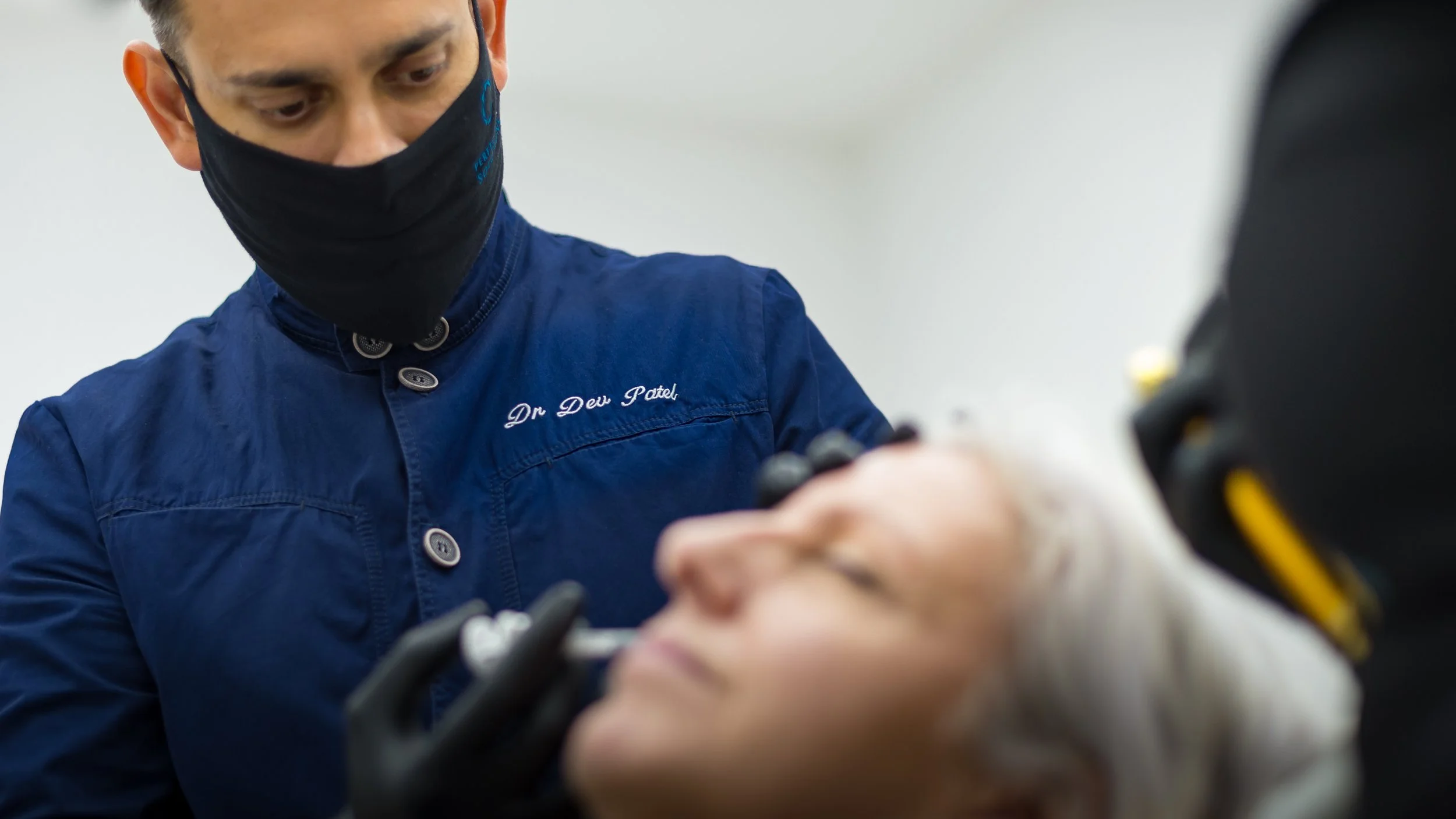 A man wearing a black face mask and blue shirt that has 'Dr. Dev Patel' embroidered on it, appears to be performing a medical or dental procedure, focusing intently on the task.