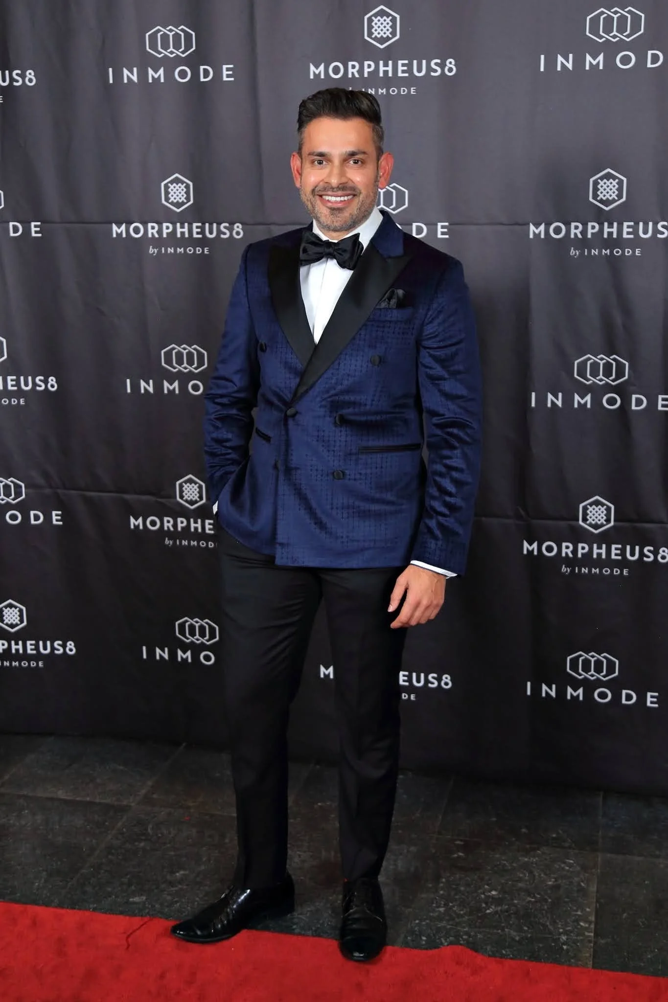 A man in a tuxedo standing on a red carpet in front of a black backdrop with logos for INMODE and MORPHEUS8, smiling at the camera.