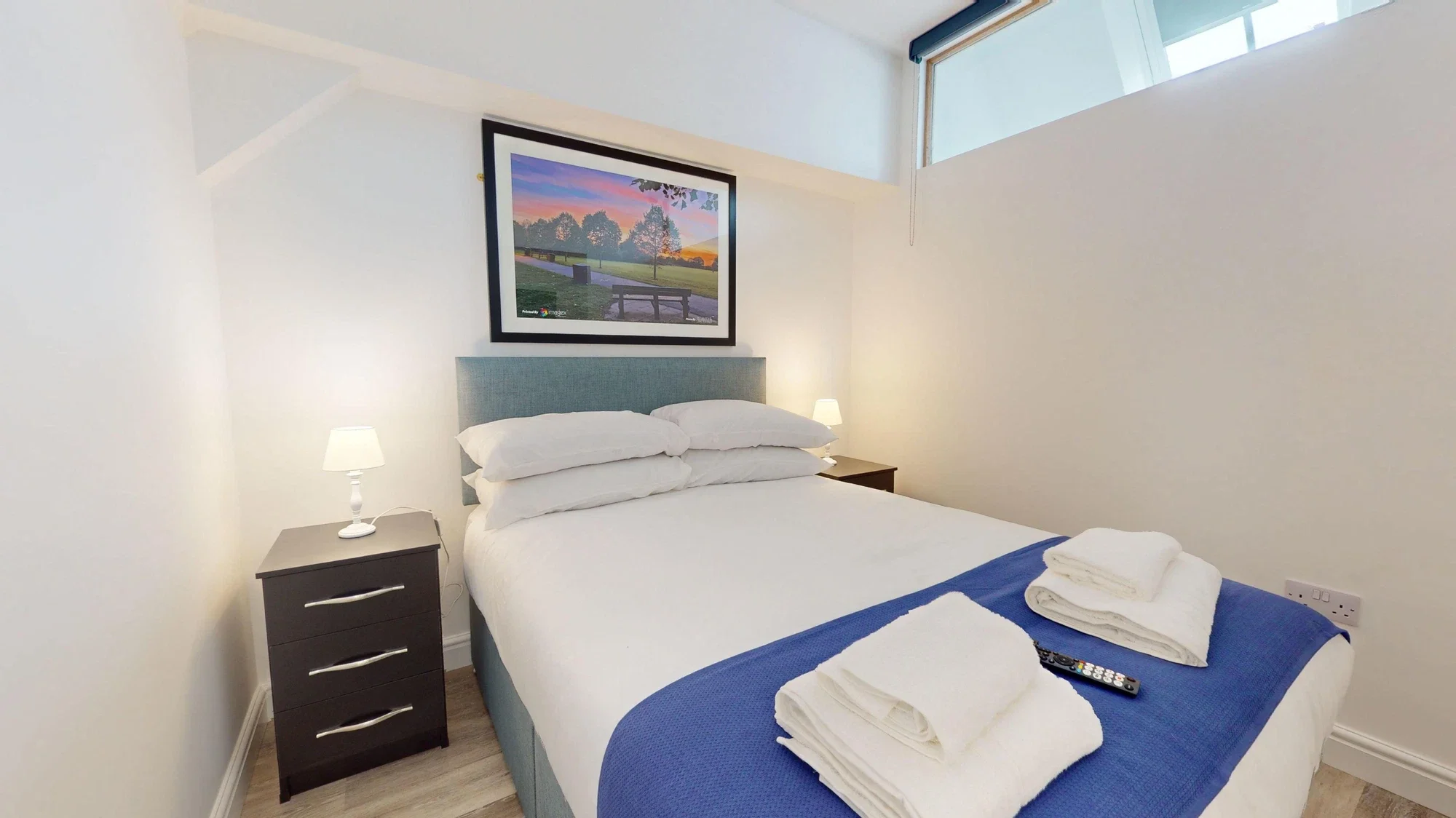 A bedroom with a double bed, white bedding, and four pillows. There are two small bedside tables with lamps, a framed landscape picture on the wall, and neatly folded towels and a remote control on the bed. The room has white walls and a small high window near the ceiling.