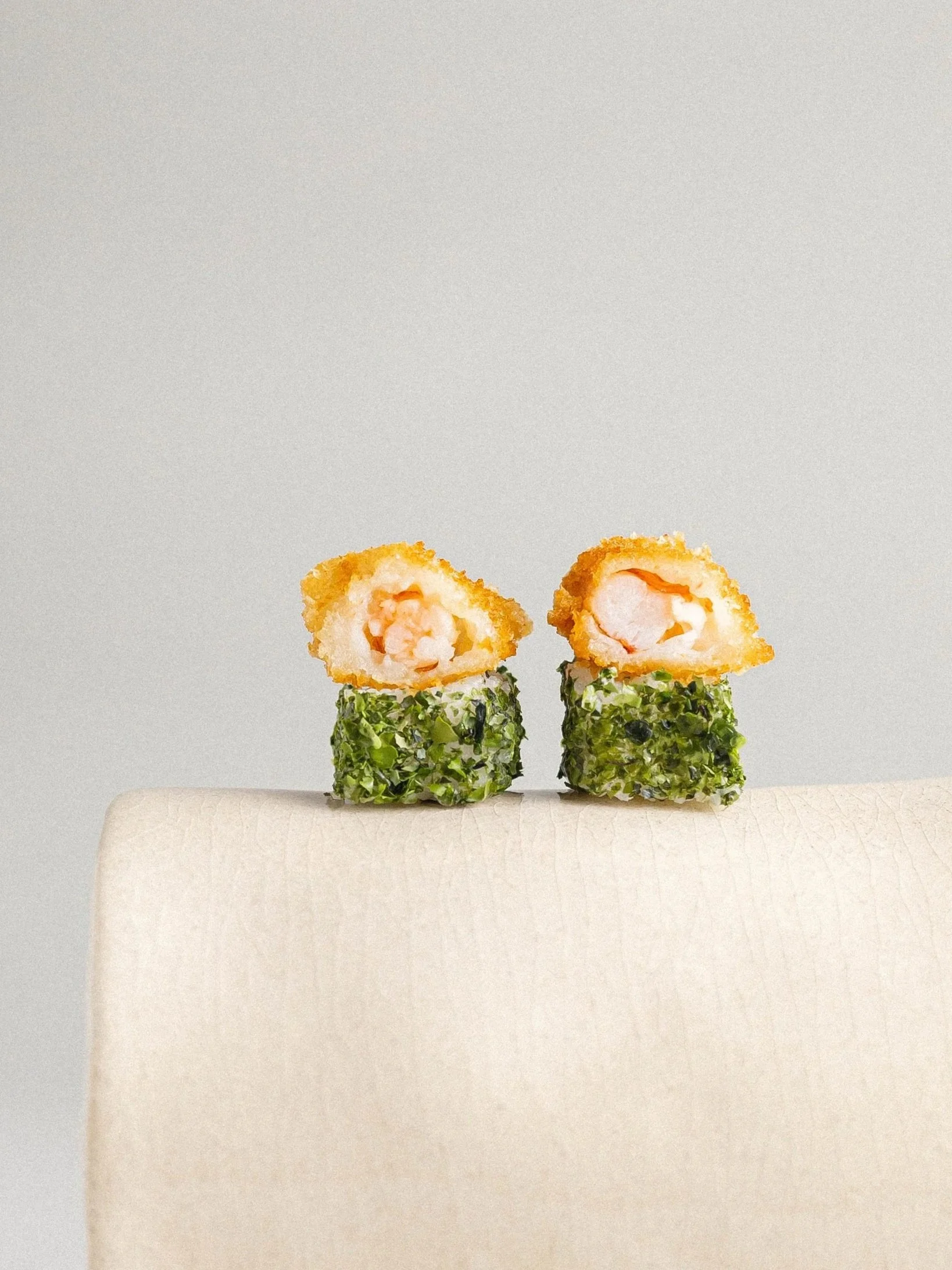 Close-up of two sushi rolls with green seaweed and white rice, topped with fried shrimp tempura pieces, on a white textured surface.