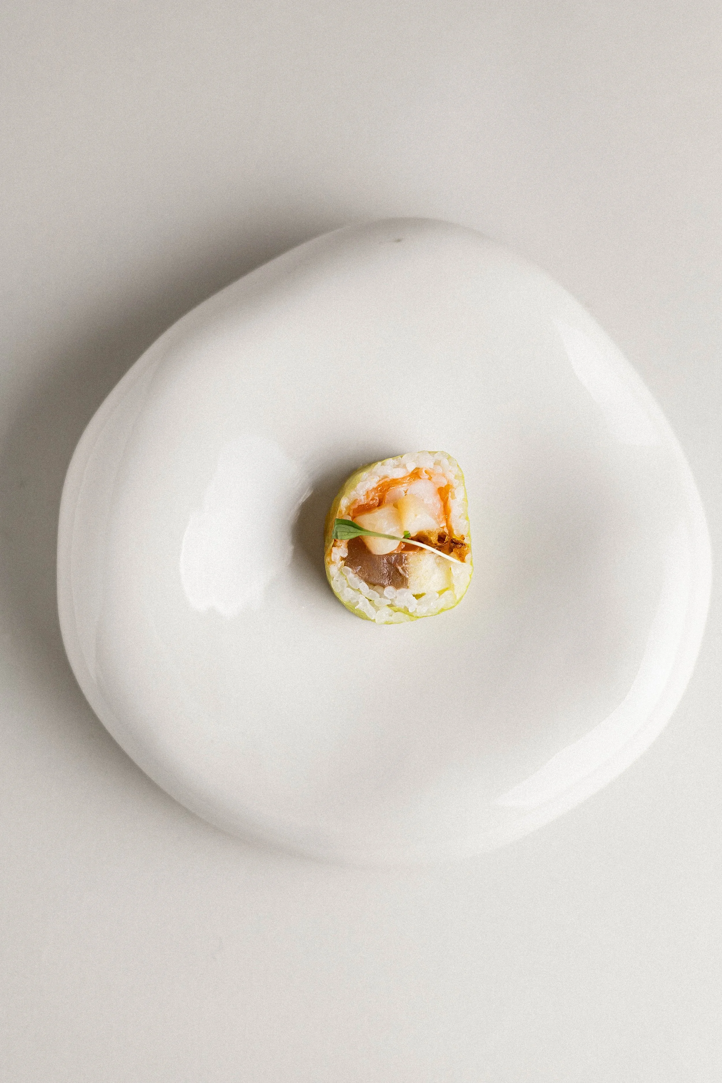 A single piece of sushi on a plain white plate, featuring rice, fish, and a small green garnish.