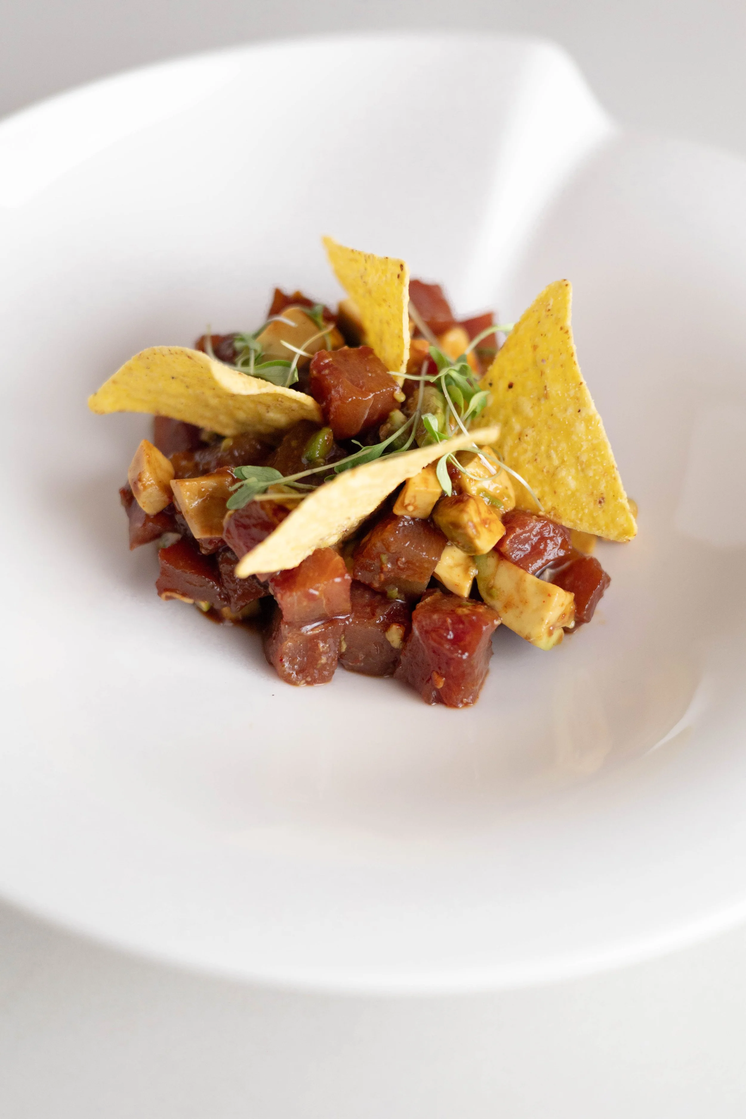 Close-up of a small portion of hash with diced meat, topped with microgreens and three yellow tortilla chips, served on a white plate.