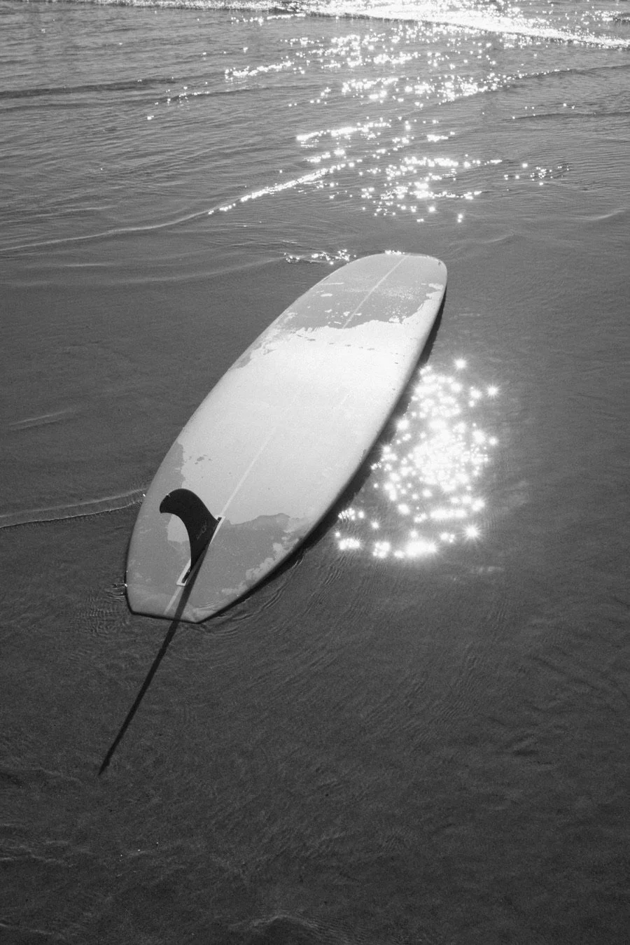 A surfboard lying upside down in the water near the shore with the sun reflecting off the surface.