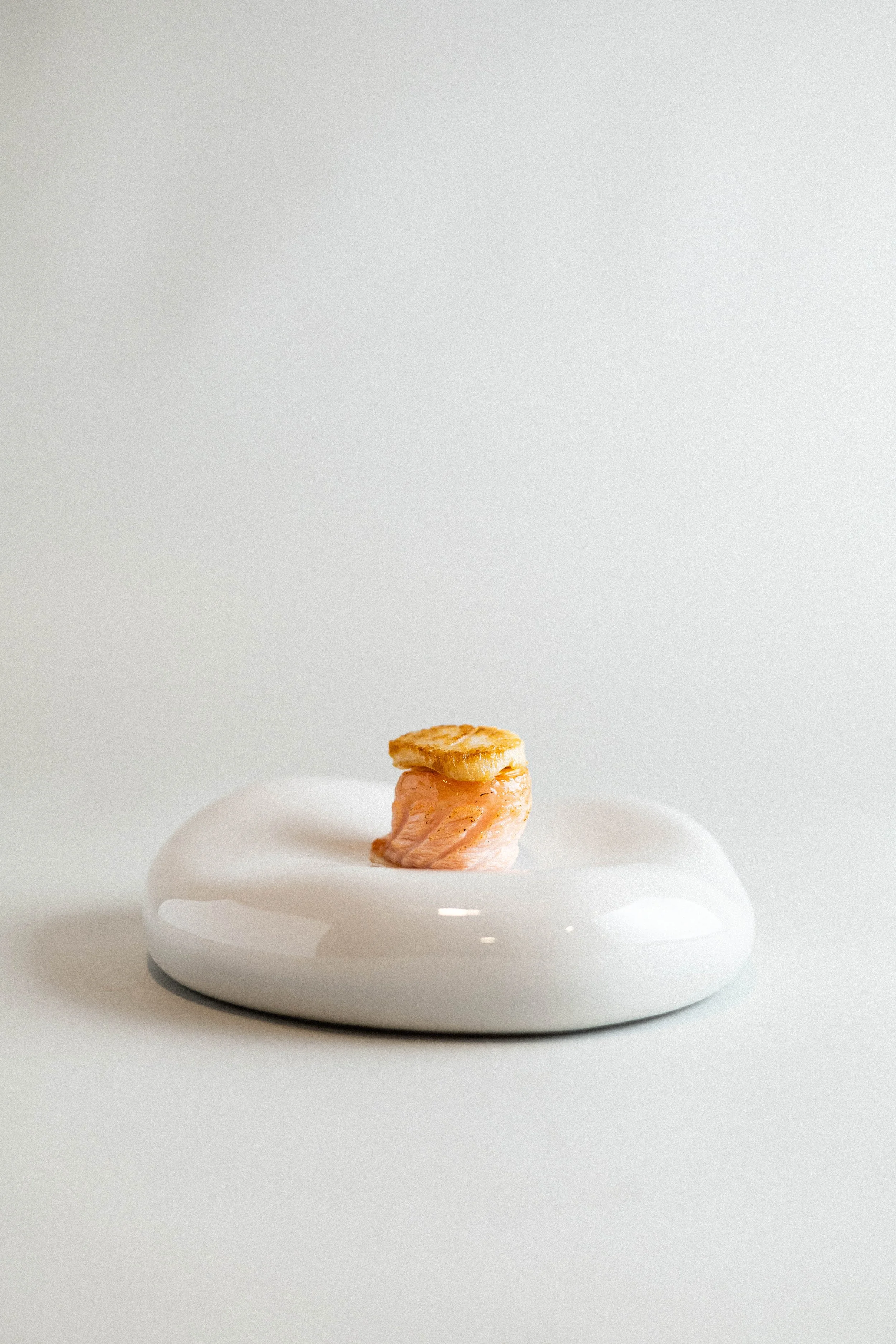 A small piece of baked salmon with a slice of roasted plantain on top, placed on a white, modern, ceramic plate with a minimalist design.