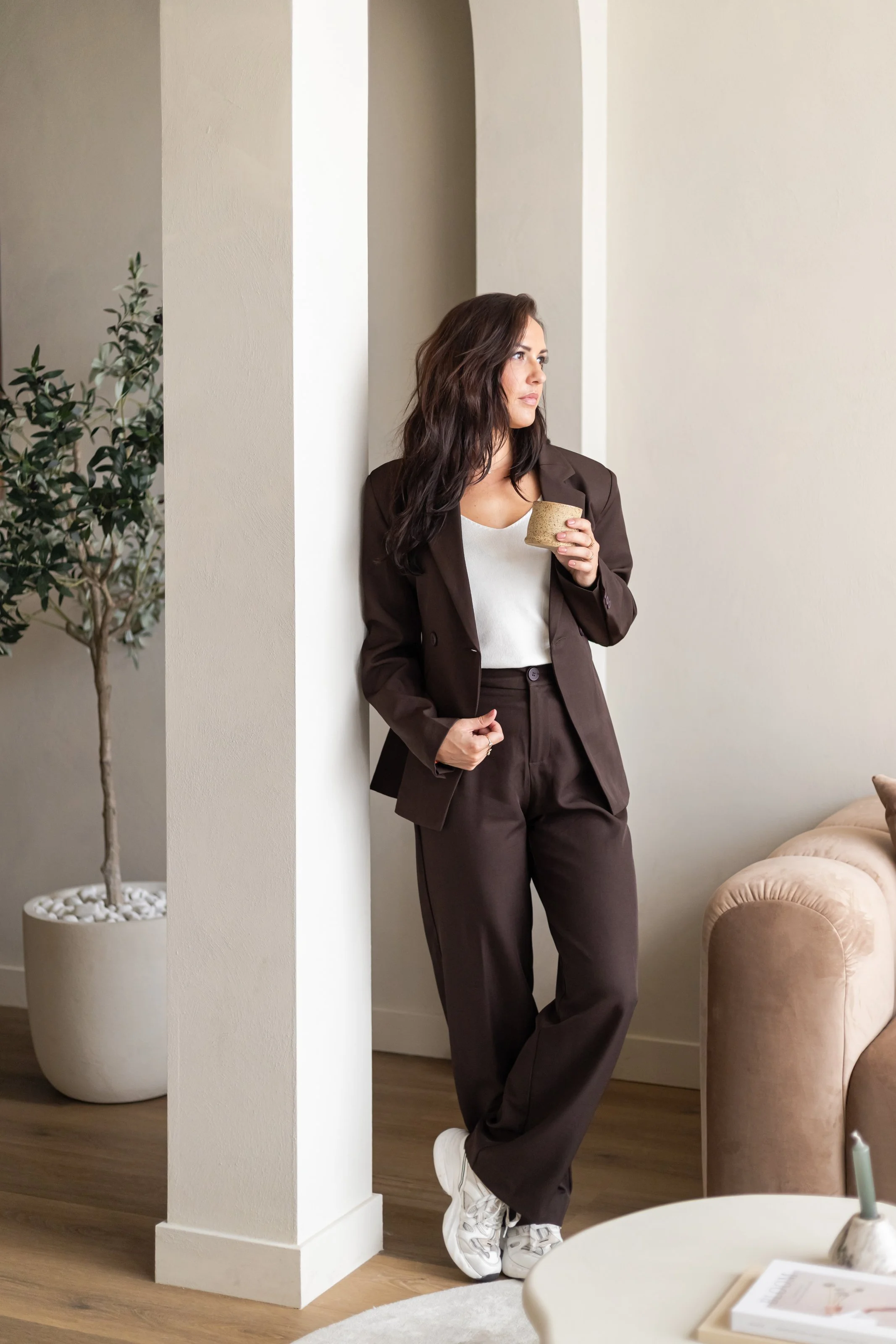 Een vrouw in een bruine pak met witte sneakers leunt tegen een witmuur, terwijl ze een kopje vasthoudt en naar rechts kijkt in een interieur met een beige bank en een plantenpot.