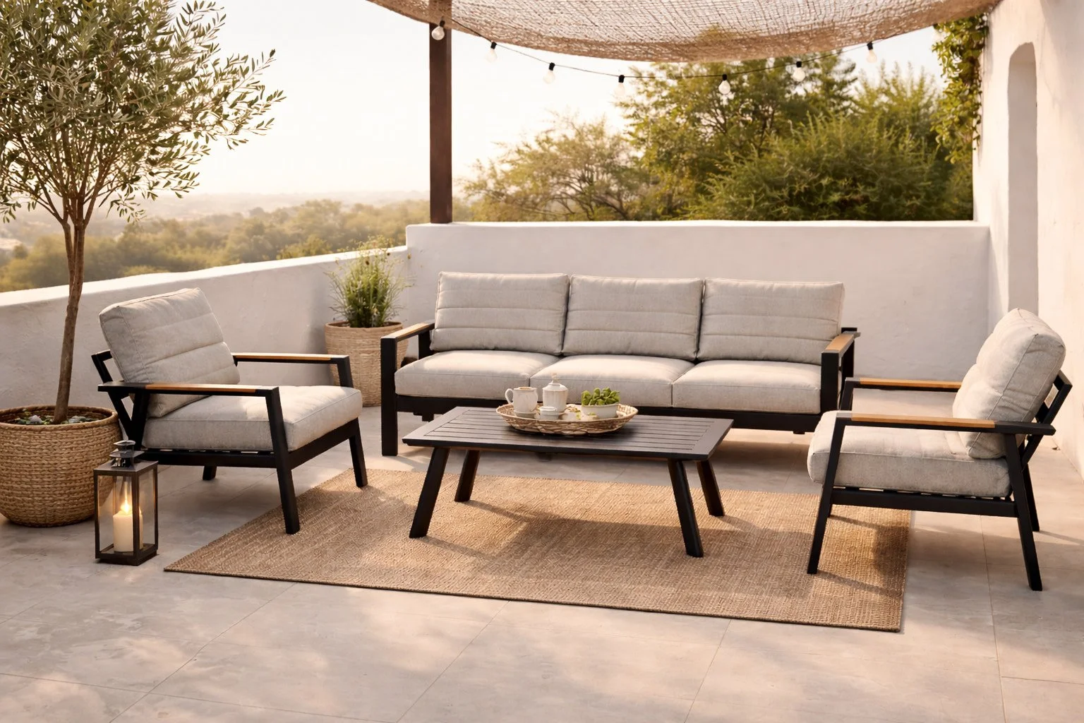 Een terras met wit en zwart meubilair, een bank en twee fauteuils, en een lage tafel met een dienblad met kopjes en een plant. Er staan twee grote potplanten en een lantaarn met een kaars. Een overkapping met lampjes en een muur maken het terras gezellig en uitnodigend.