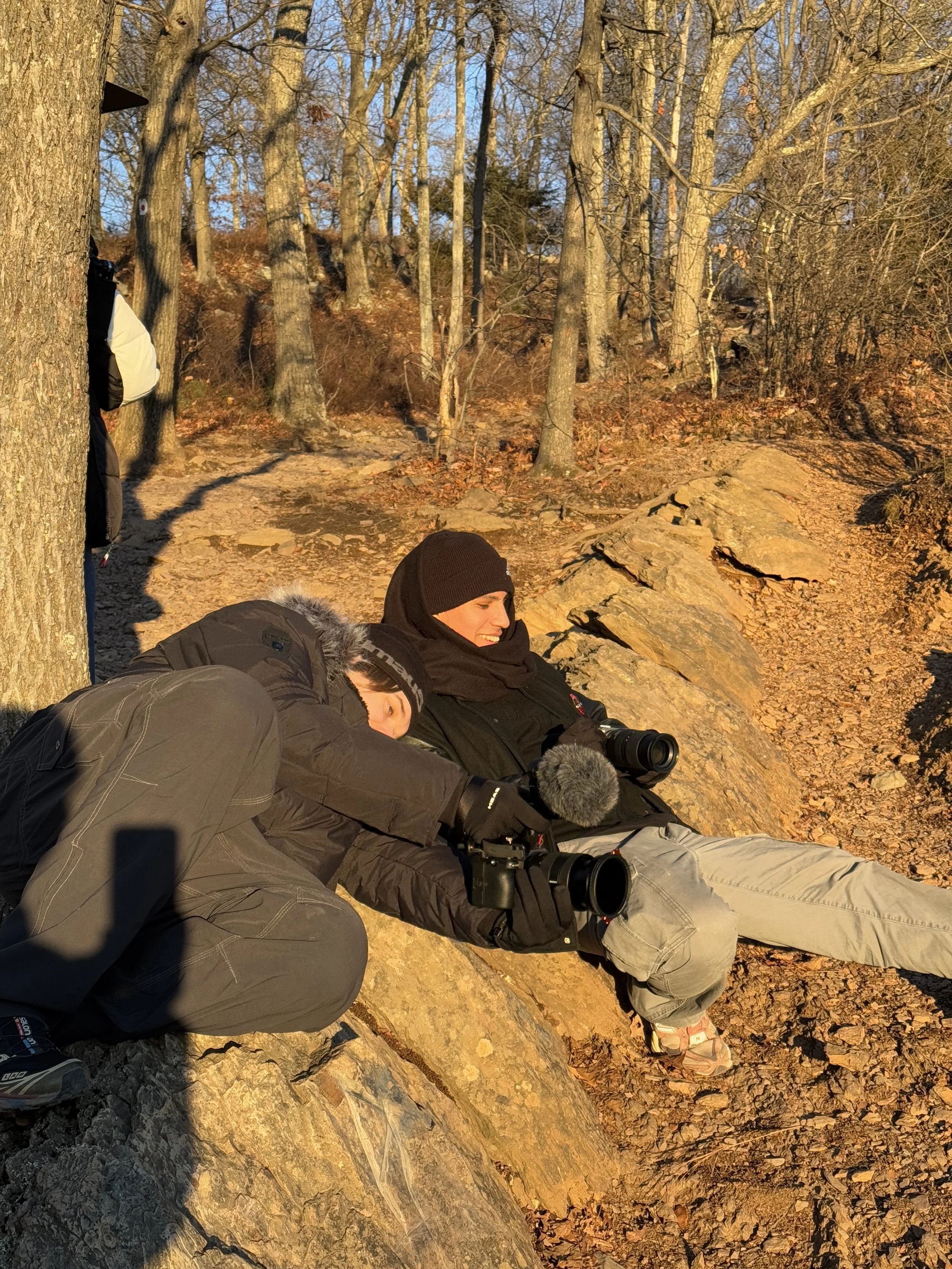 Two people sitting and lying on rocks in a wooded area during sunset, both holding cameras with microphones and both dressed warmly for cold weather.