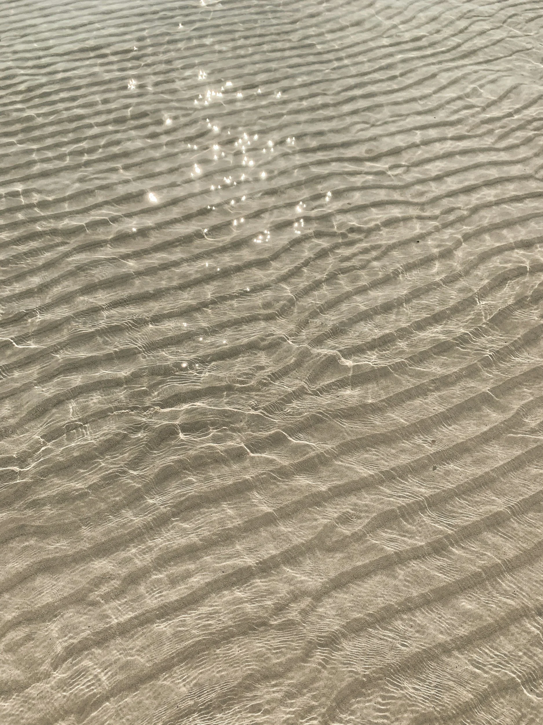 Close-up of clear shallow water on a sandy beach with ripples and sunlight reflections.