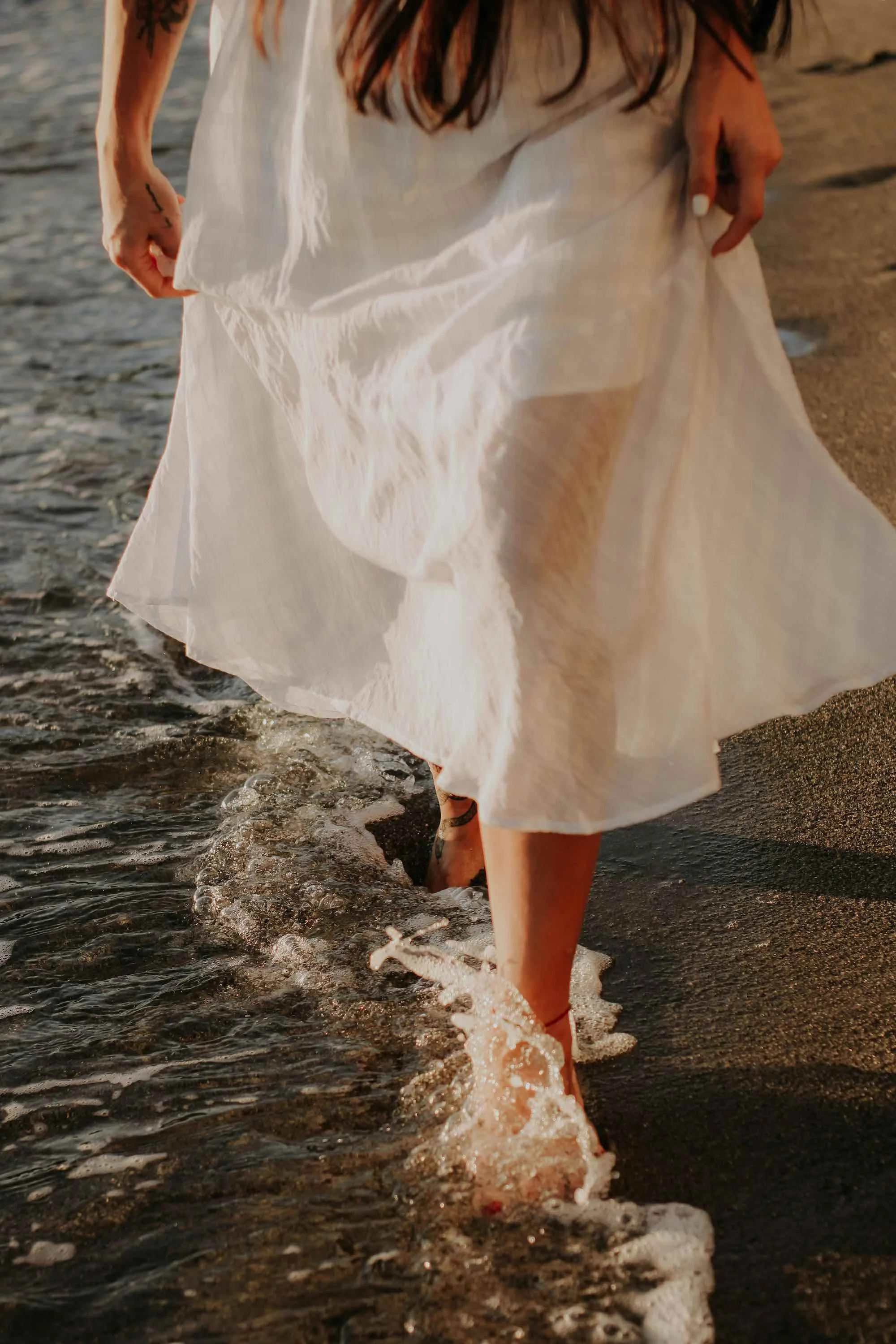 Person walking at the beach, wearing a white dress, with one hand holding the dress and the other hand resting on the thigh, as the waves roll in around their feet.