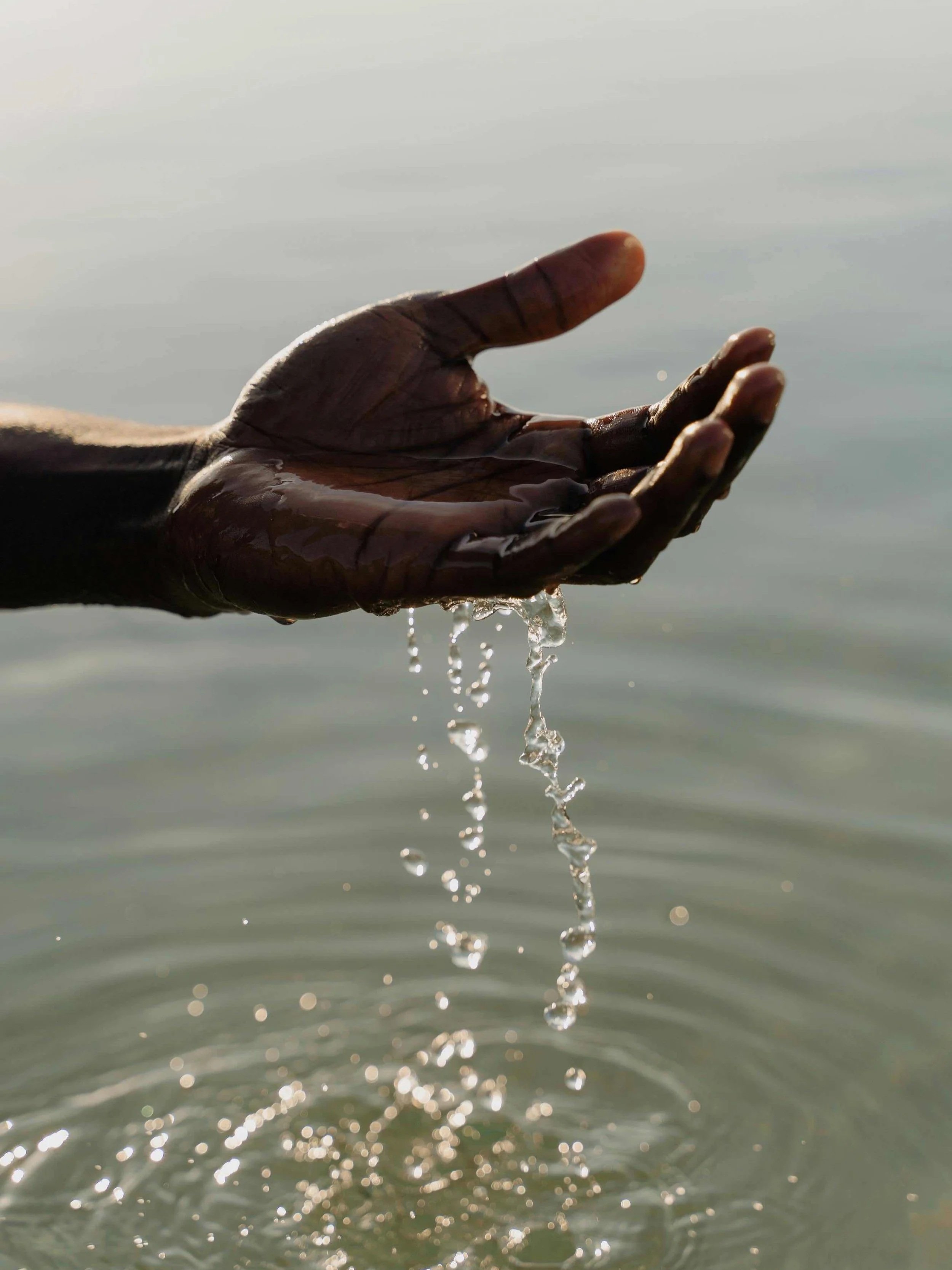 A person with dark skin is holding water in their hand over a body of water, with water dripping from their fingers.