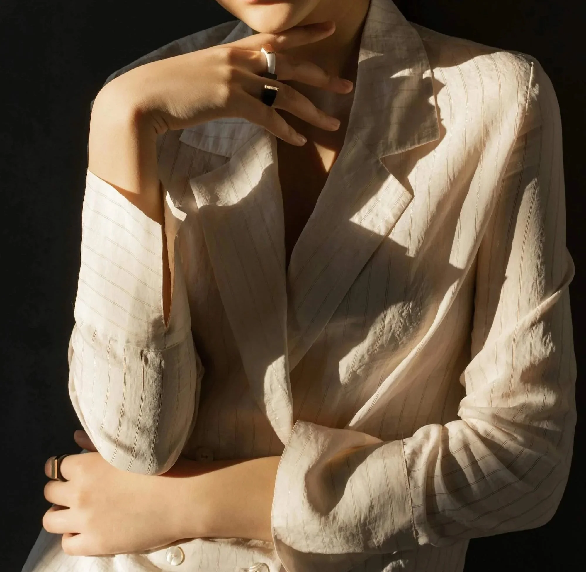 A person wearing a light-colored, striped shirt with the collar open, resting their chin on their hand, with a dark background.