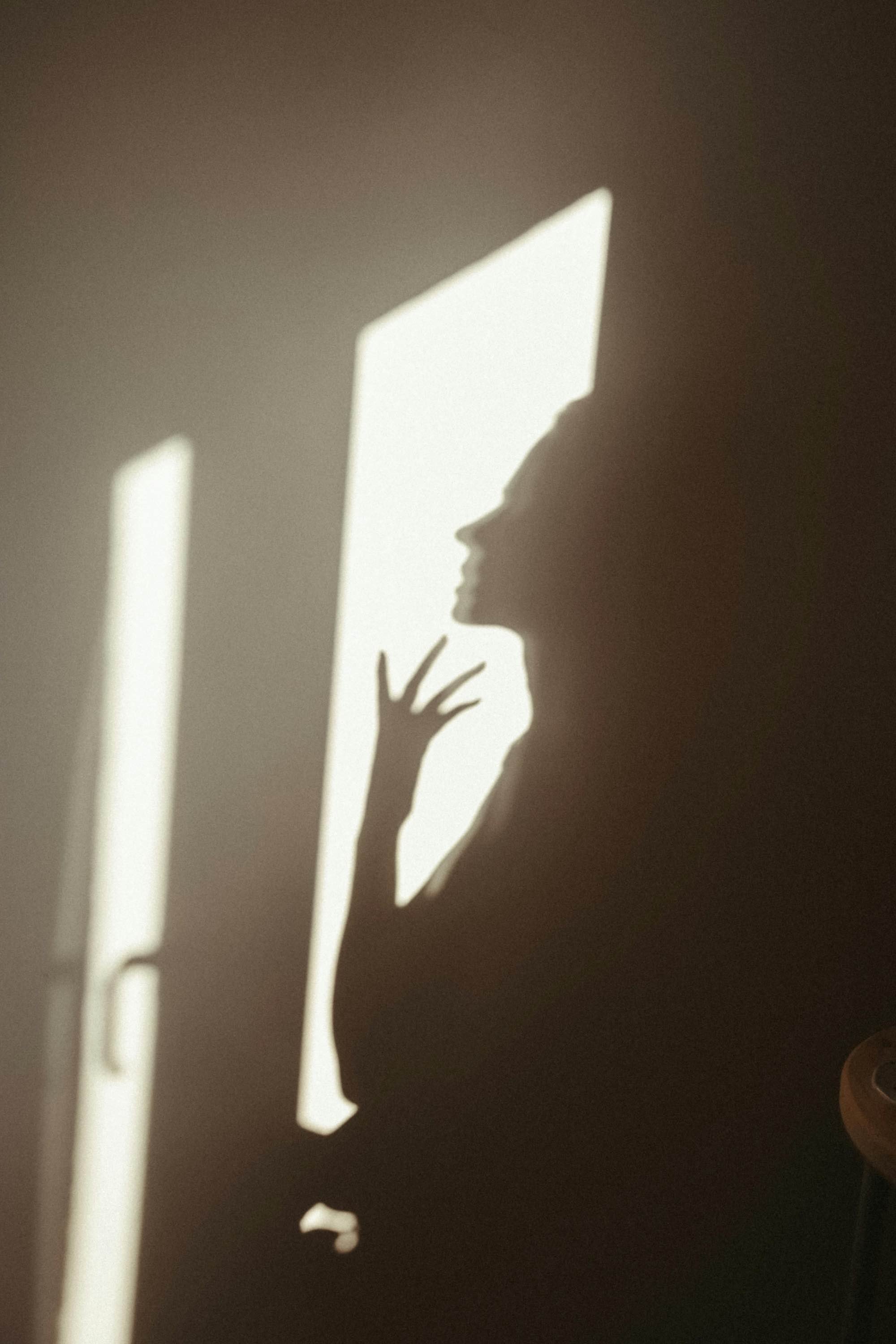 Shadow of a woman with long hair, touching her face, cast on a wall by sunlight through a window.