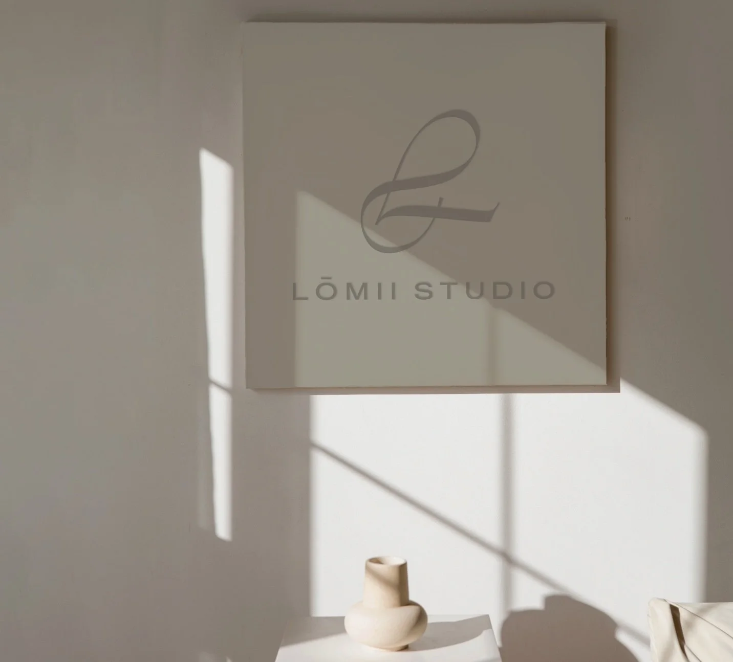 Artwork with a logo and the text 'LõMII STUDIO' on a white wall with natural sunlight casting shadows, and a small white ceramic vase on a table below.