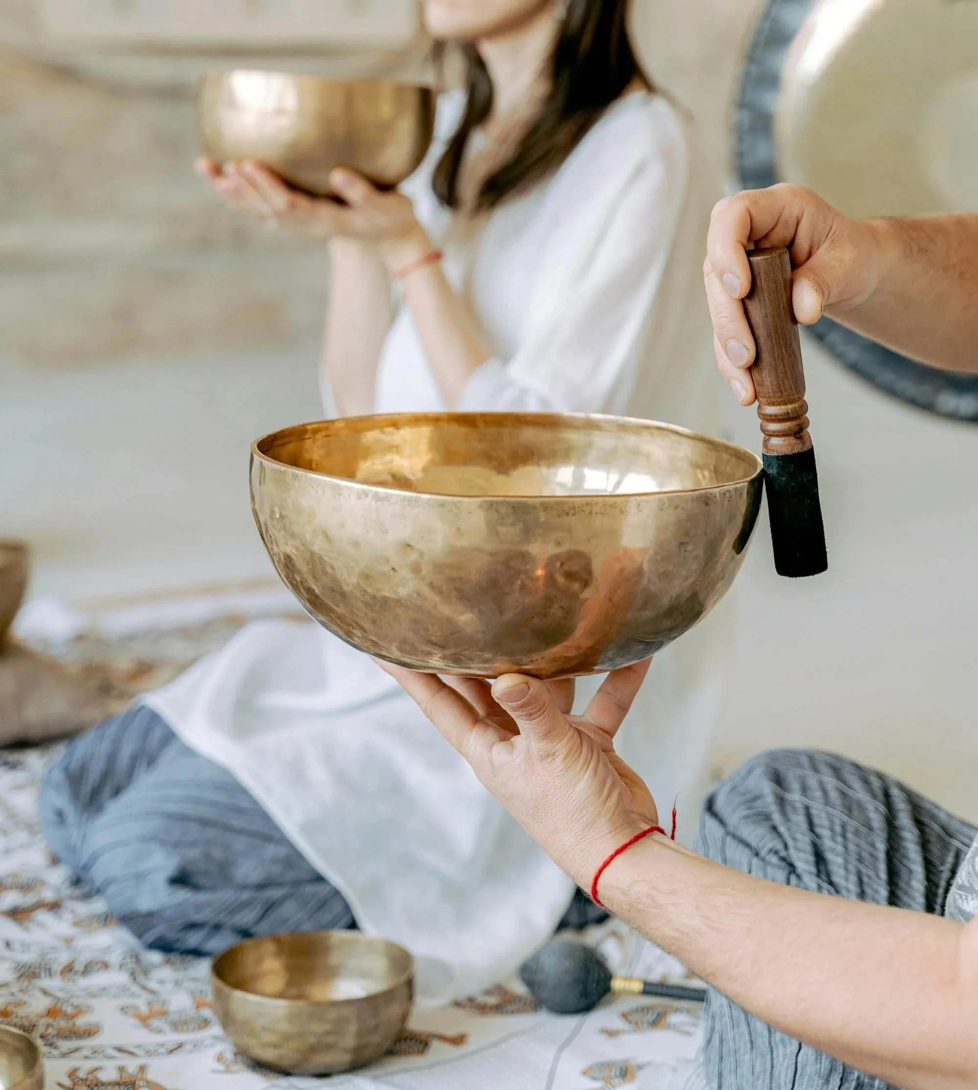 Two people hold metal singing bowls, with one person striking a bowl with a mallet and the other holding a bowl near their chest. They are sitting on a patterned cloth with additional bowls around them.