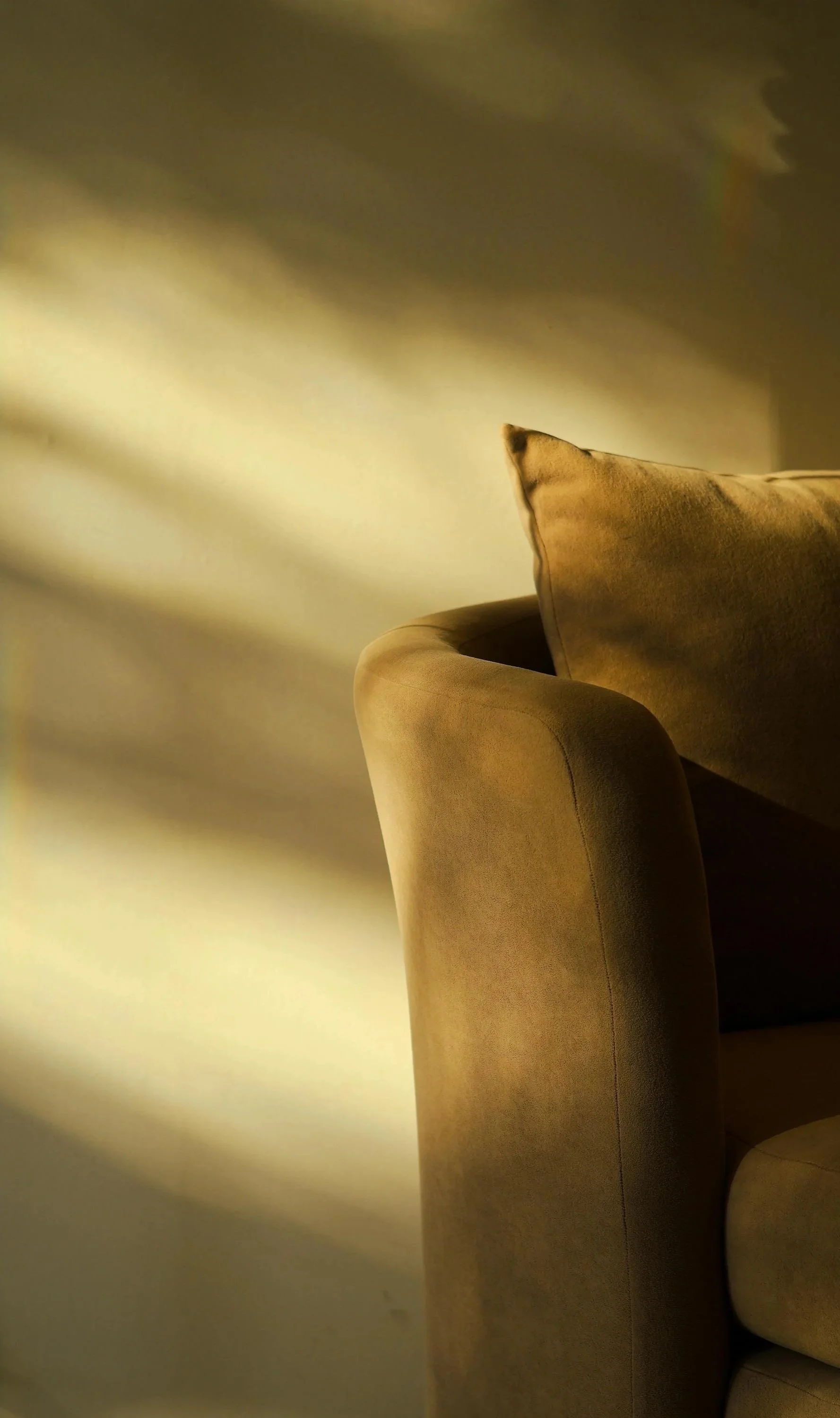 Close-up of a beige sofa with some throw pillows, with warm sunlight casting shadows on the wall behind it.