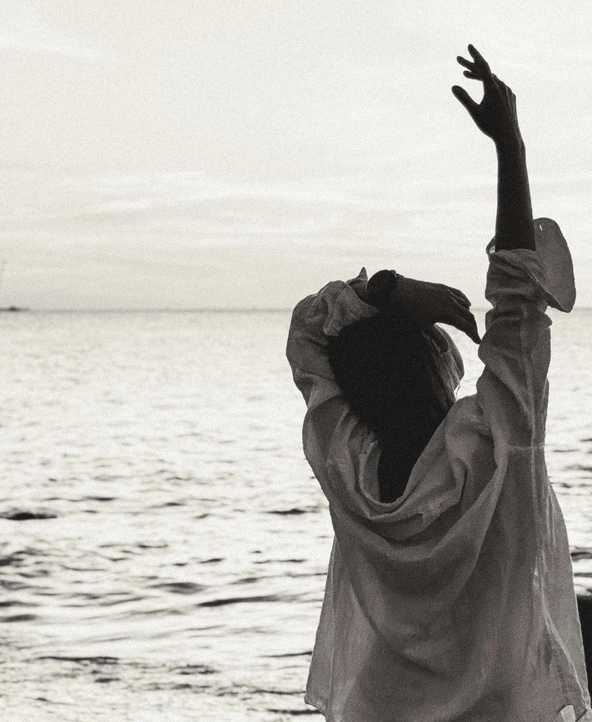 A person standing on the beach near water, wearing a hoodie, with one hand raised and the other holding their head, facing the water during sunset or sunrise.