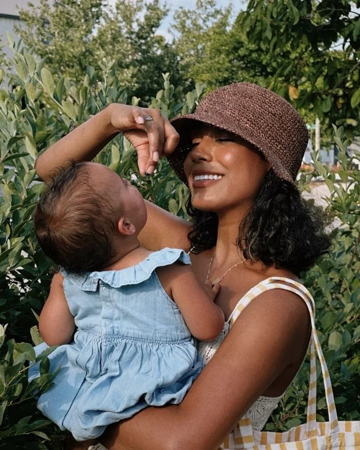 A woman wearing a brown sunhat holding a baby girl in her arms outdoors among greenery, both smiling.