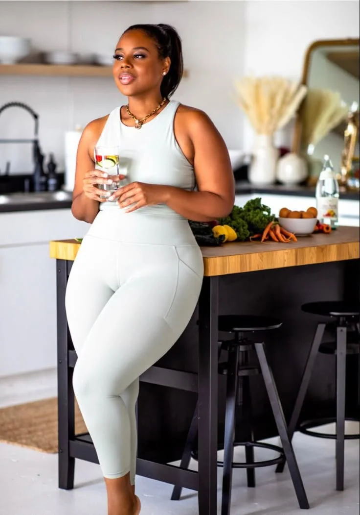 A woman in white workout clothes stands in a modern kitchen, holding a glass of water, with fresh vegetables on the counter beside her.