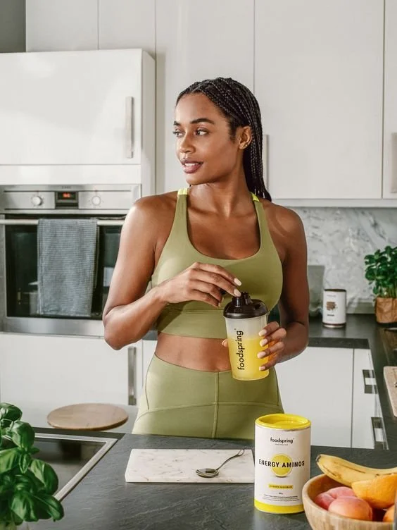 A woman in green athletic wear preparing a drink in a modern kitchen with white cabinets, a black countertop, and a container of energy amino acids on the counter.